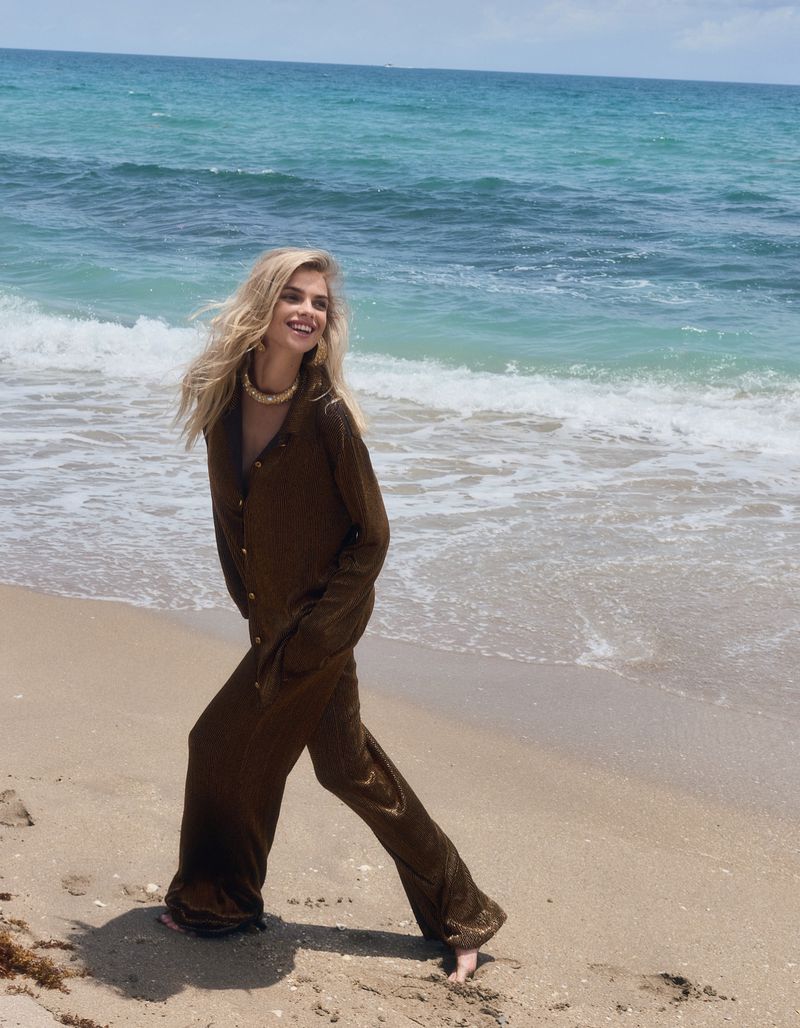 A blonde woman in brown pajamas walks barefoot on a sandy beach next to ocean waves on a sunny day, looking off to the side.