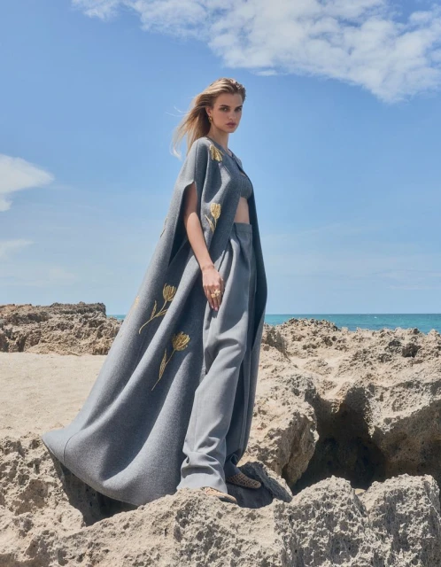 A blonde model stands on coastal rocks in a grey wool cape and pants with floral detail against a blue sky and ocean backdrop.