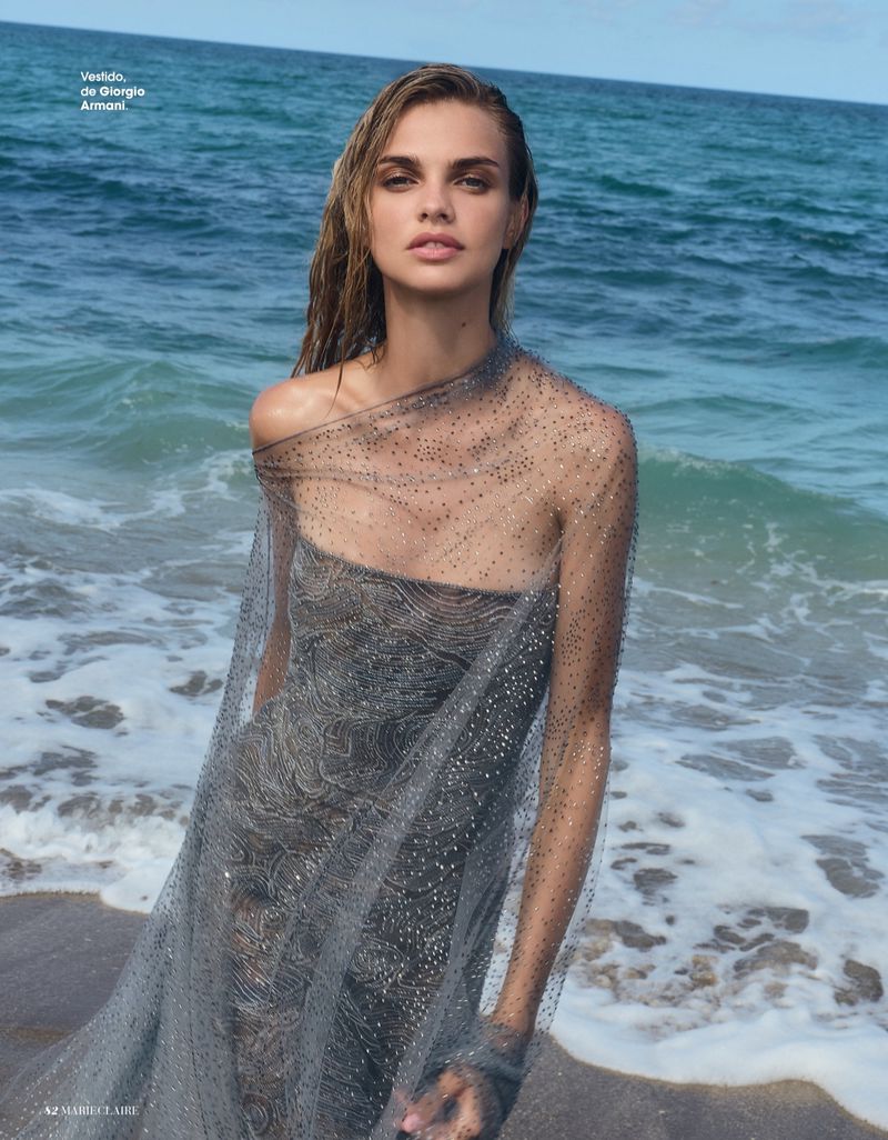 Model Dasha Khlystun wearing a sheer embellished grey gown on a sandy shoreline with breaking ocean waves.