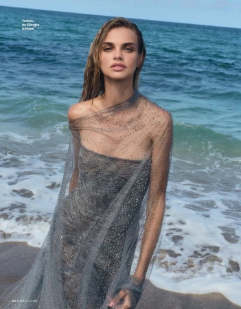 Model Dasha Khlystun wearing a sheer embellished grey gown on a sandy shoreline with breaking ocean waves.