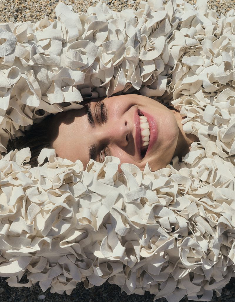 Close-up of a smiling woman's face surrounded by textured, cream-colored fabric, laying on a sandy, pebble-strewn surface outdoors.