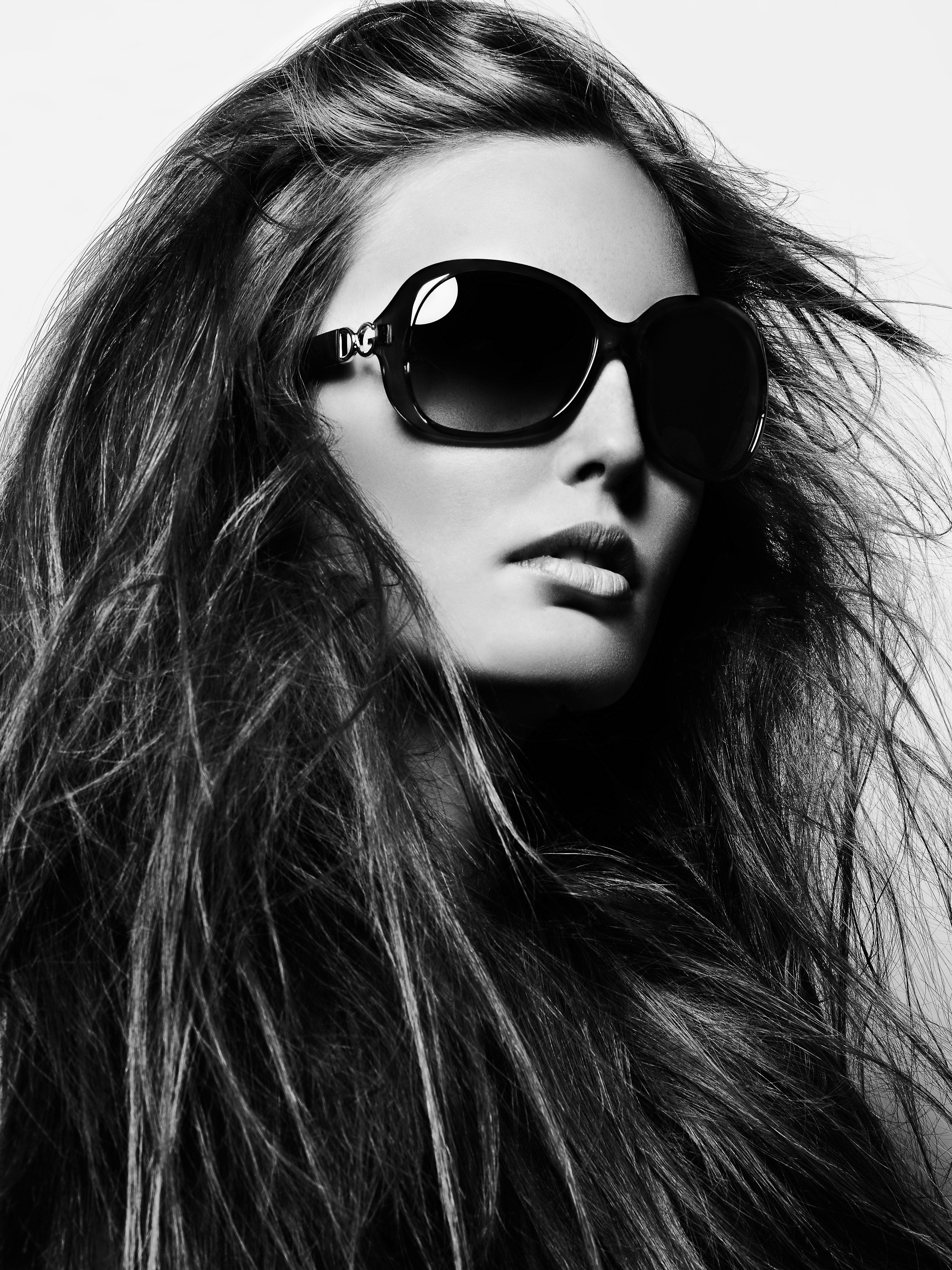 Black and white headshot of an adult woman with long, flowing hair wearing large black sunglasses against a bright white backdrop.