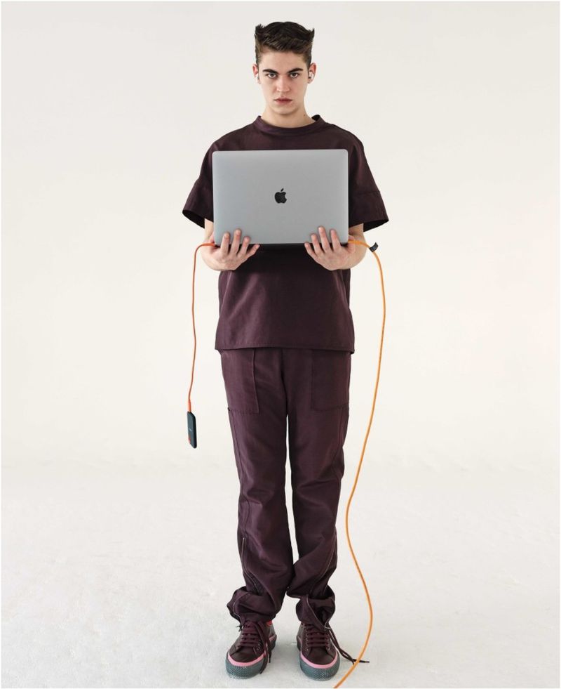 Full-shot of a young adult man with a serious facial expression holding a gray laptop, wearing a brown monochrome outfit in a white studio.