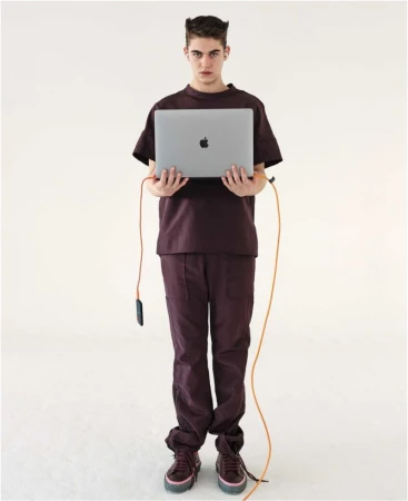 Full-shot of a young adult man with a serious facial expression holding a gray laptop, wearing a brown monochrome outfit in a white studio.