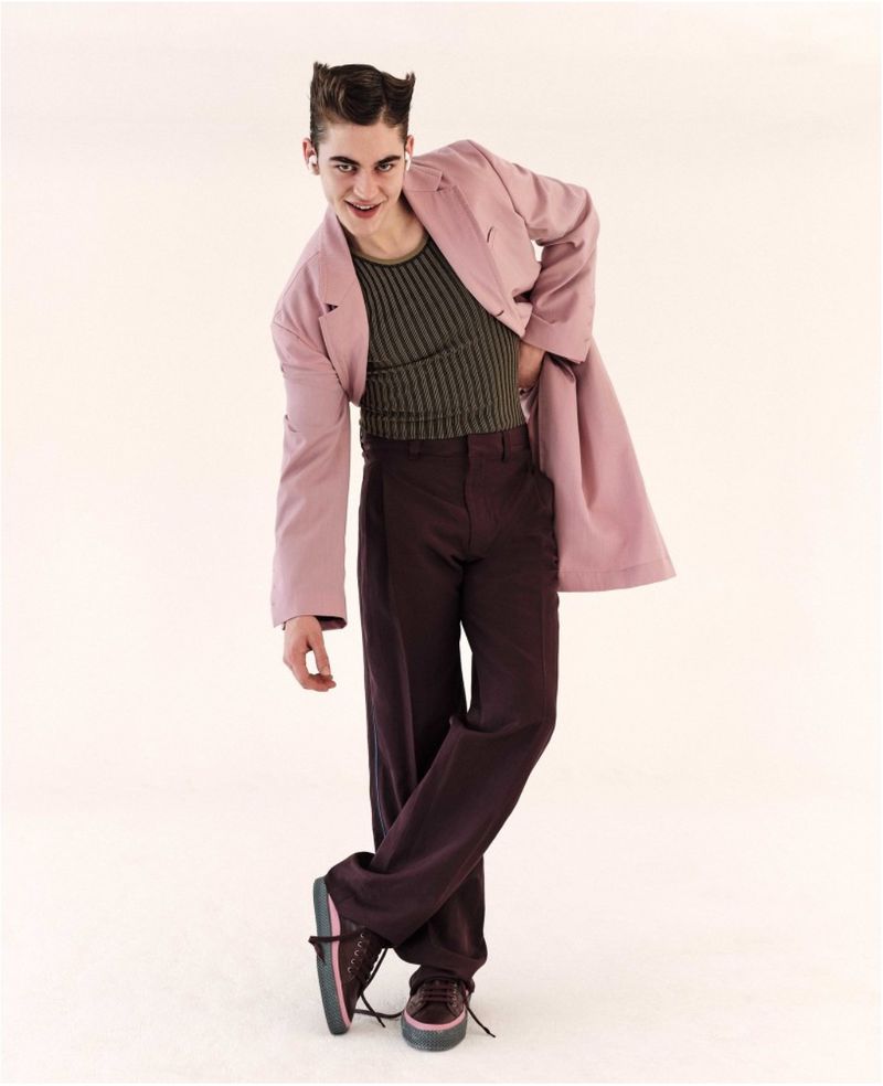 A stylish adult male posing in a pink jacket and brown pants, standing with crossed legs against a neutral background in a fashion shot.