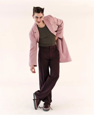 A stylish adult male posing in a pink jacket and brown pants, standing with crossed legs against a neutral background in a fashion shot.