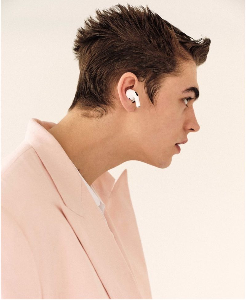Profile shot of a young adult man wearing a pink blazer and white wireless earbuds, against a clean, bright white backdrop in studio lighting.