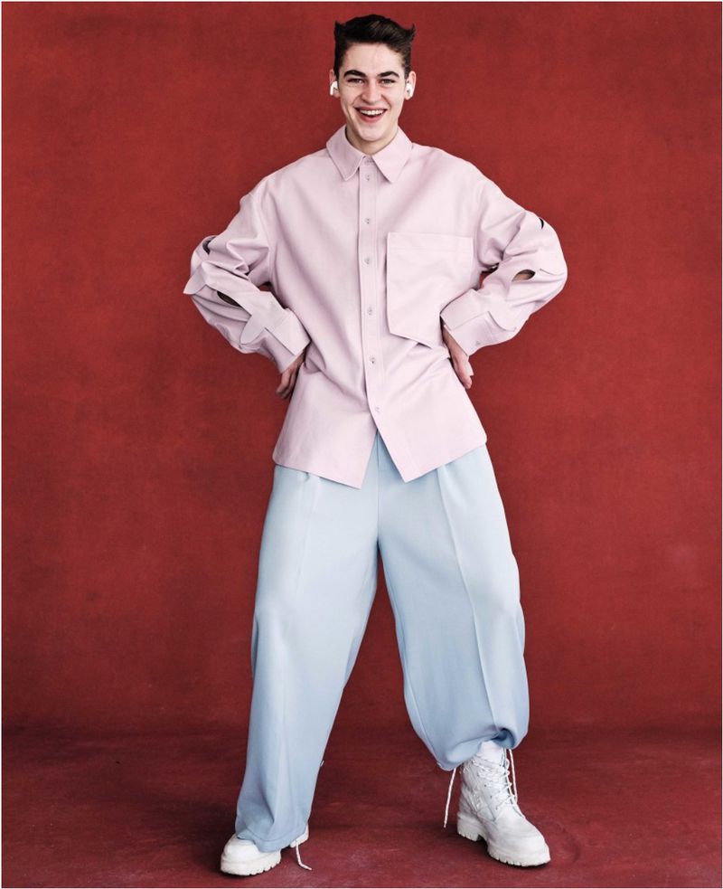 Young man poses in an oversized pink shirt and light blue pants against a red studio backdrop, highlighted by white platform boots.