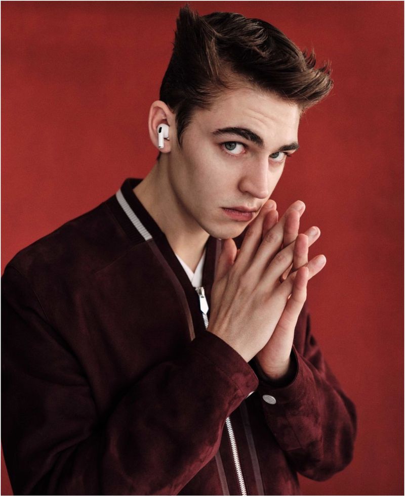 Close-up of a young adult man with light brown hair wearing a bomber jacket, posing against a solid red background.