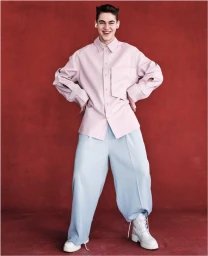 Hero Fiennes Tiffin smiling in a pink button-down shirt and light blue wide-leg trousers against a red textured wall