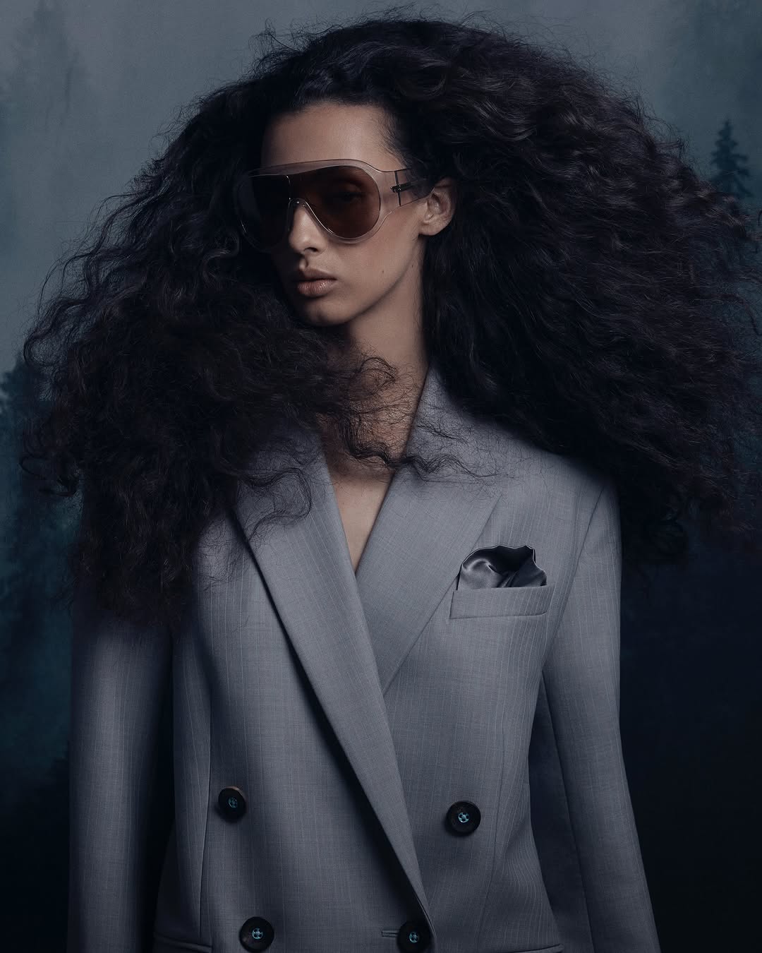 A woman with curly black hair models sunglasses and a grey blazer against a blurred forest background in a beauty fashion portrait.