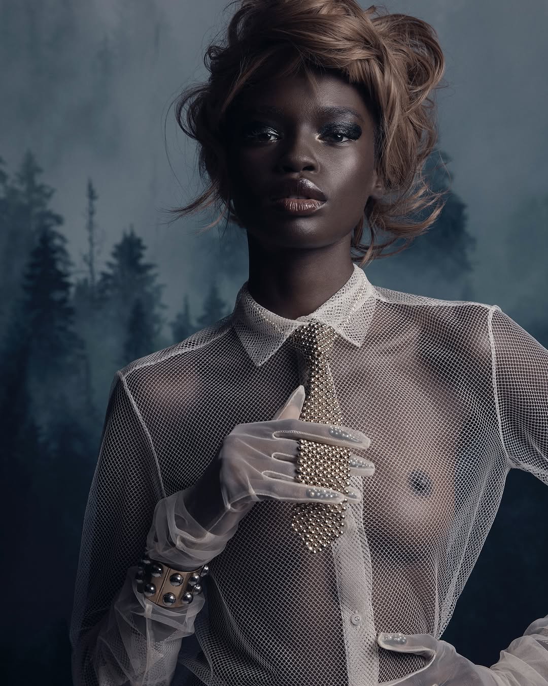 Elegant portrait of a Black woman with styled hair wearing a sheer shirt and textured tie against a smoky forest background.