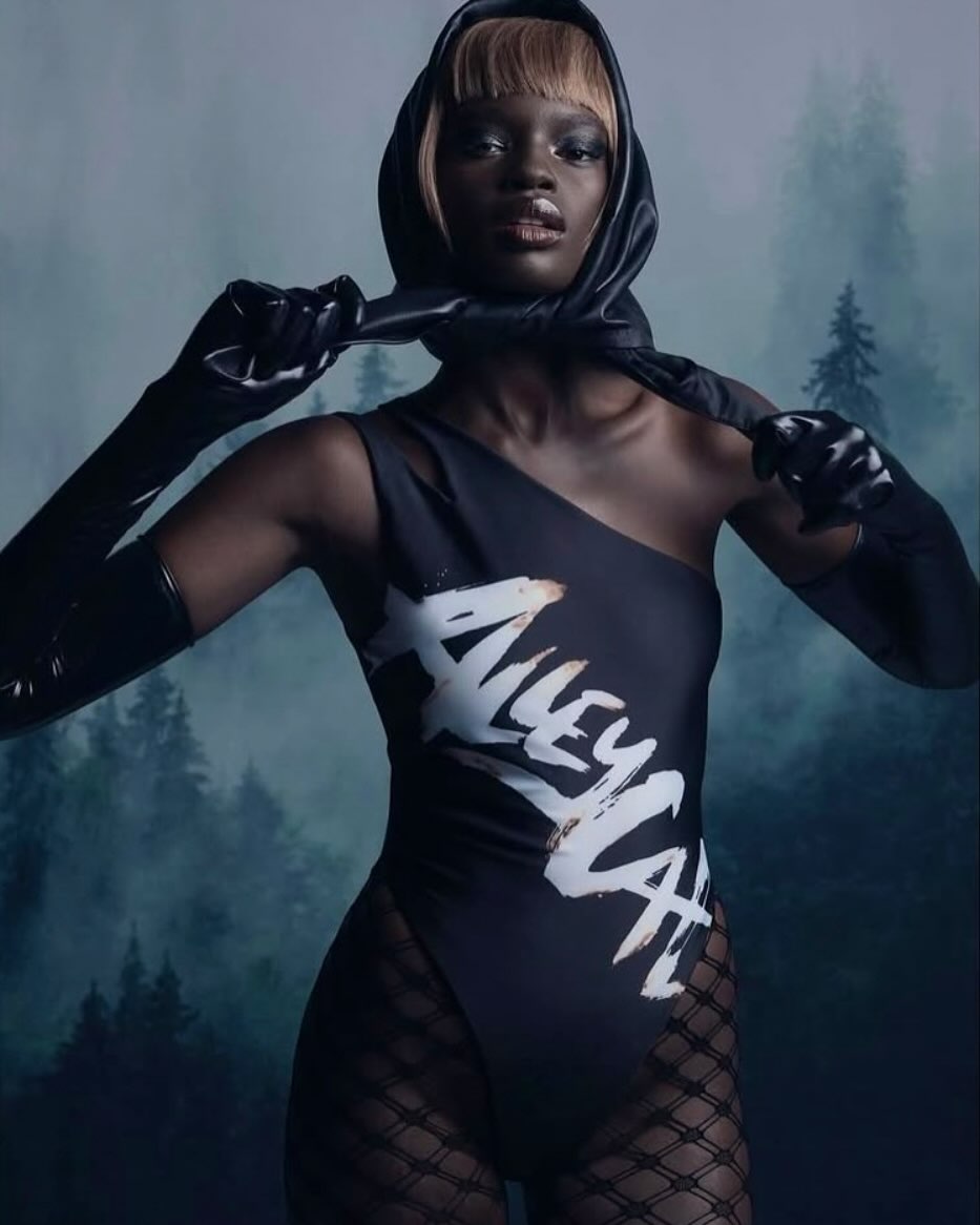 Striking fashion shot of a dark-skinned woman in a black swimsuit with white print, gloves, and headscarf, posing against a blurred forest backdrop.