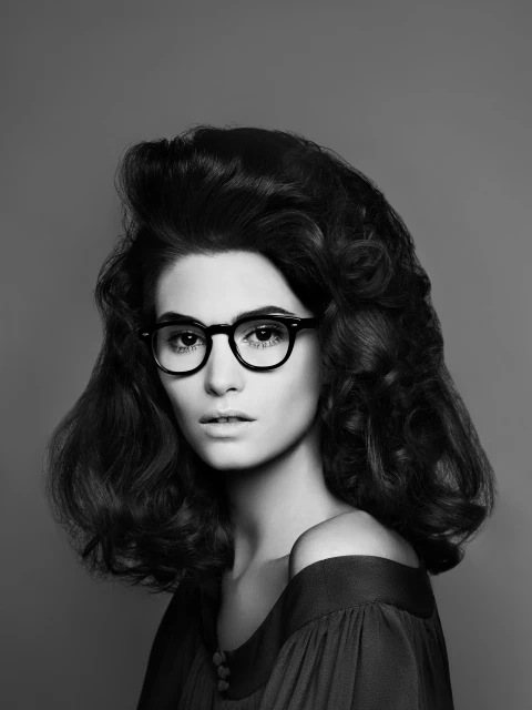 Headshot of a woman with curly dark hair, wearing black glasses and a dark off-the-shoulder top, posing in a photo studio.