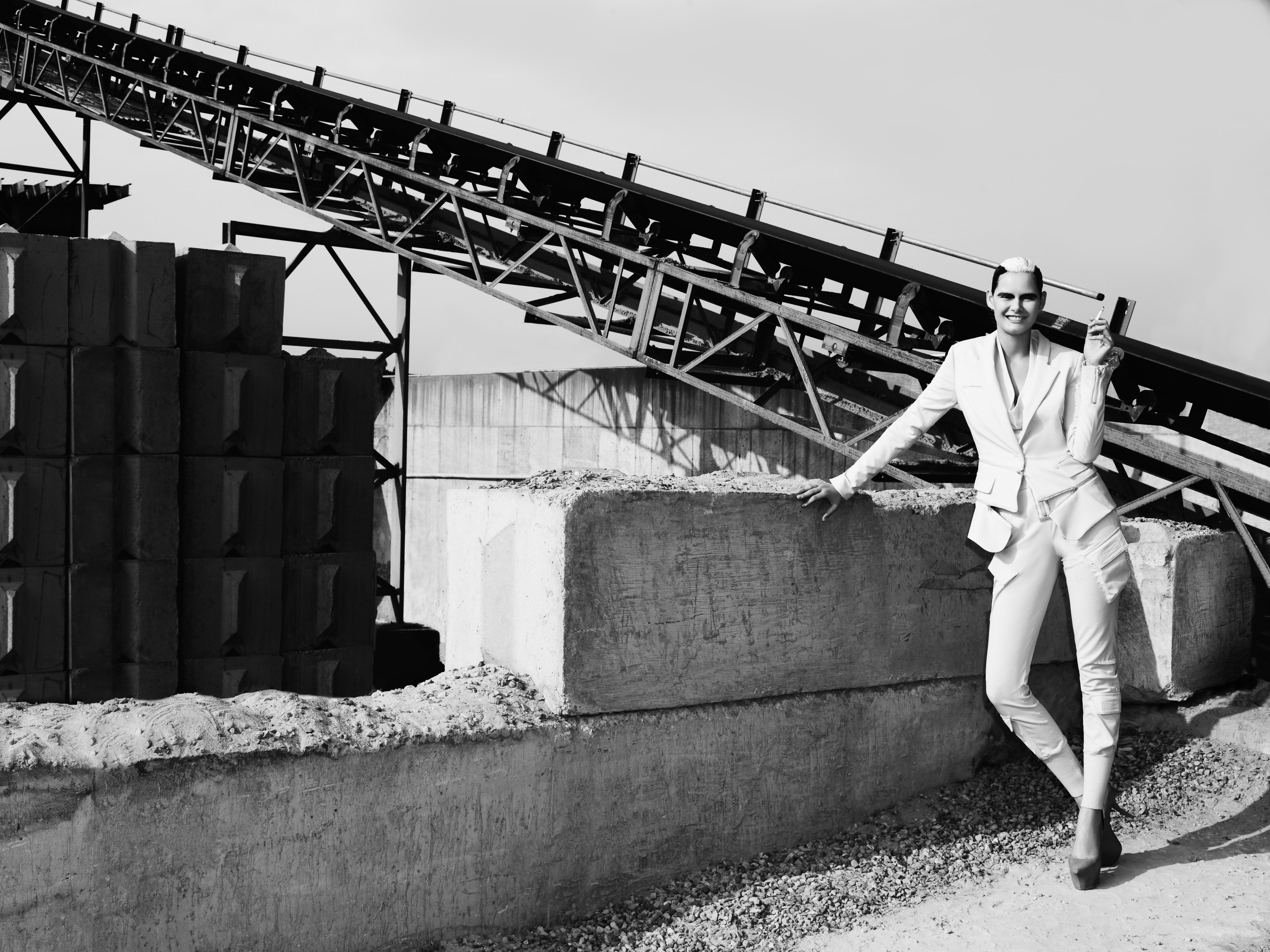 Monochrome shot of a stylish adult woman in a white suit posing by large concrete blocks, with an industrial conveyor in the background.
