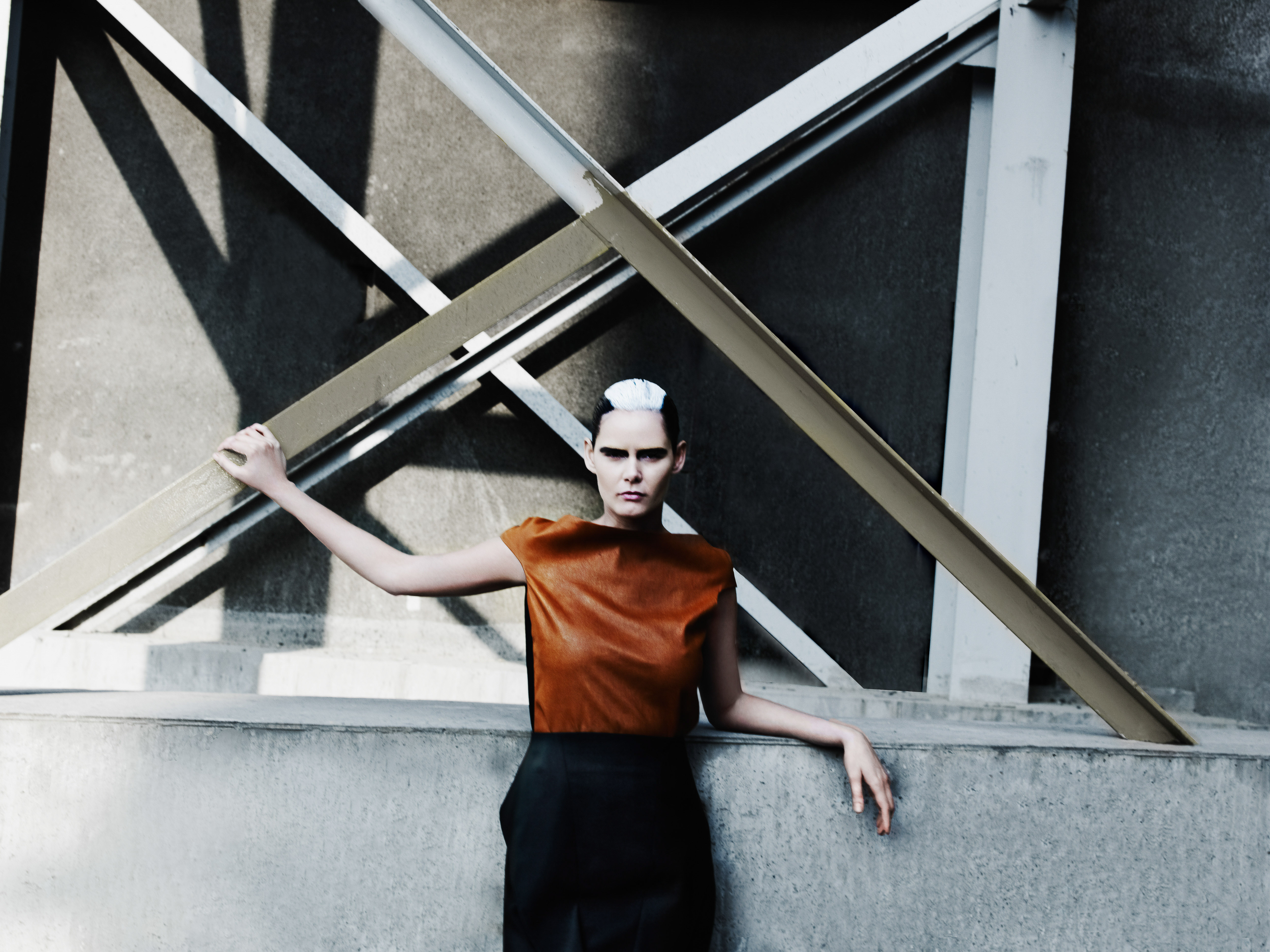 Fashion shot of an adult woman with short white and brown hair, wearing a brown top and dark skirt, posing near a concrete wall.