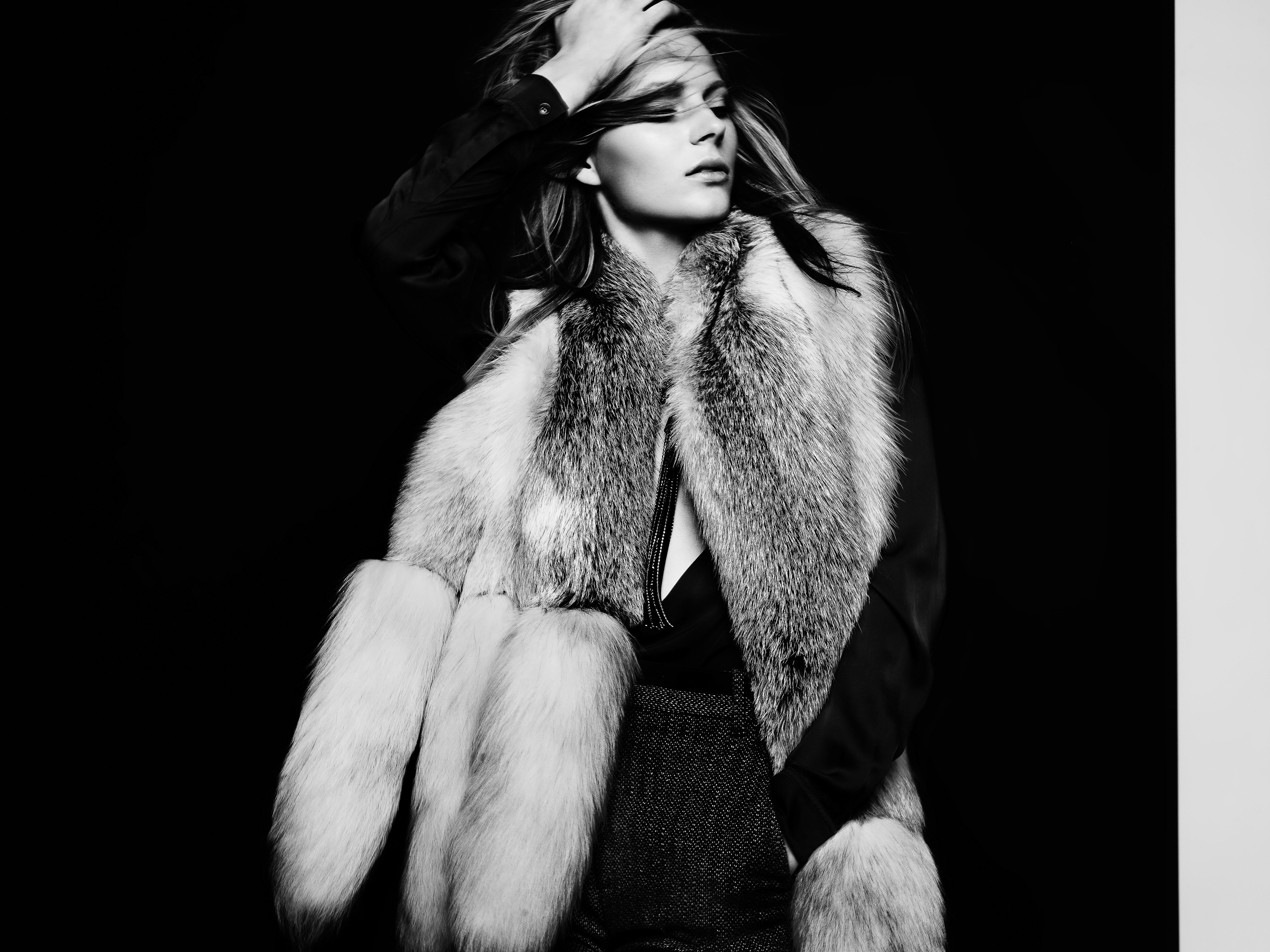 Black and white portrait of a blonde woman in a fur vest, captured in a studio setting with a dark backdrop, fashion, and luxury appeal.
