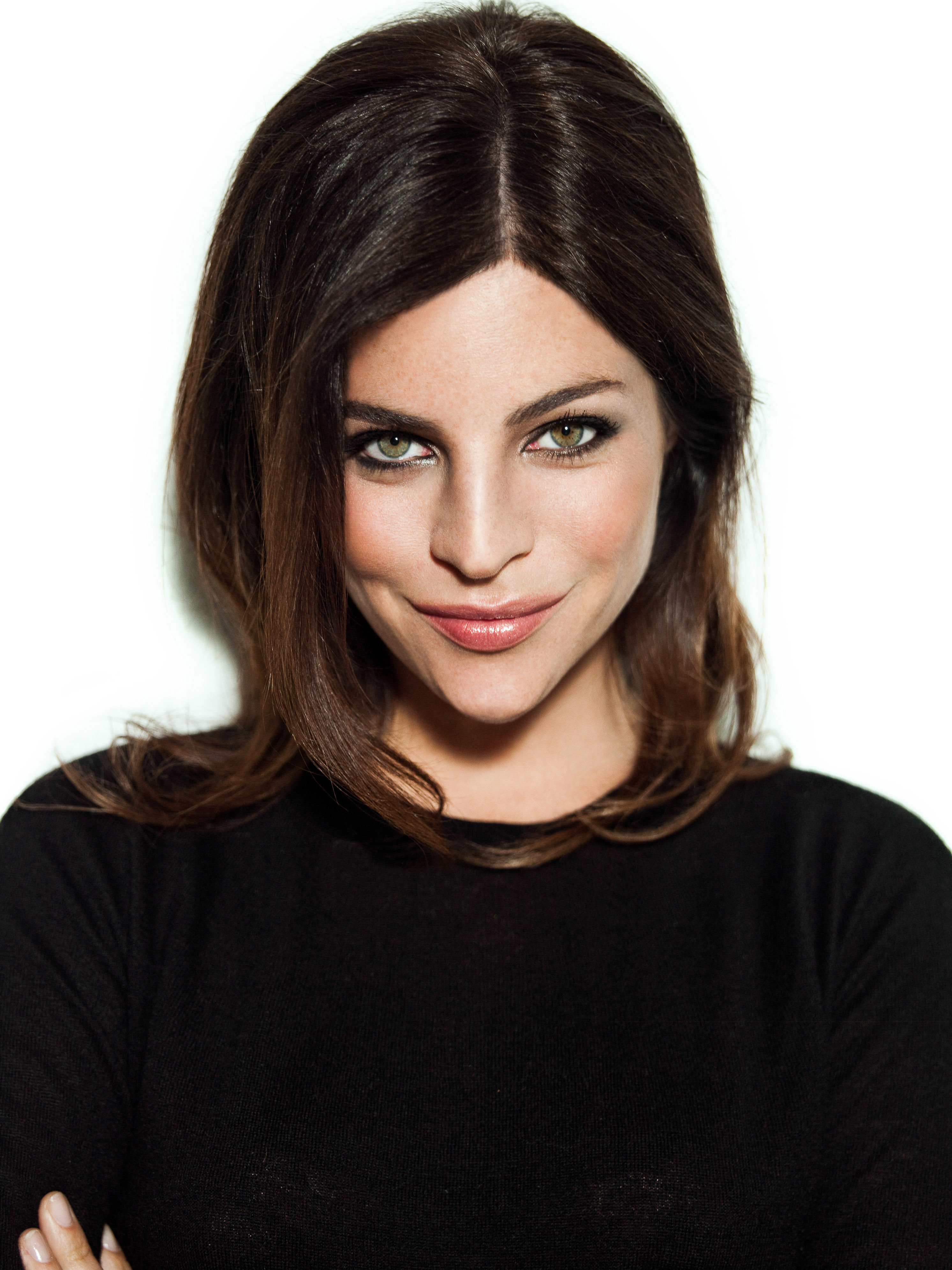 Close-up portrait of a dark-haired, green-eyed woman with a subtle smile wearing a black top against a bright white background.