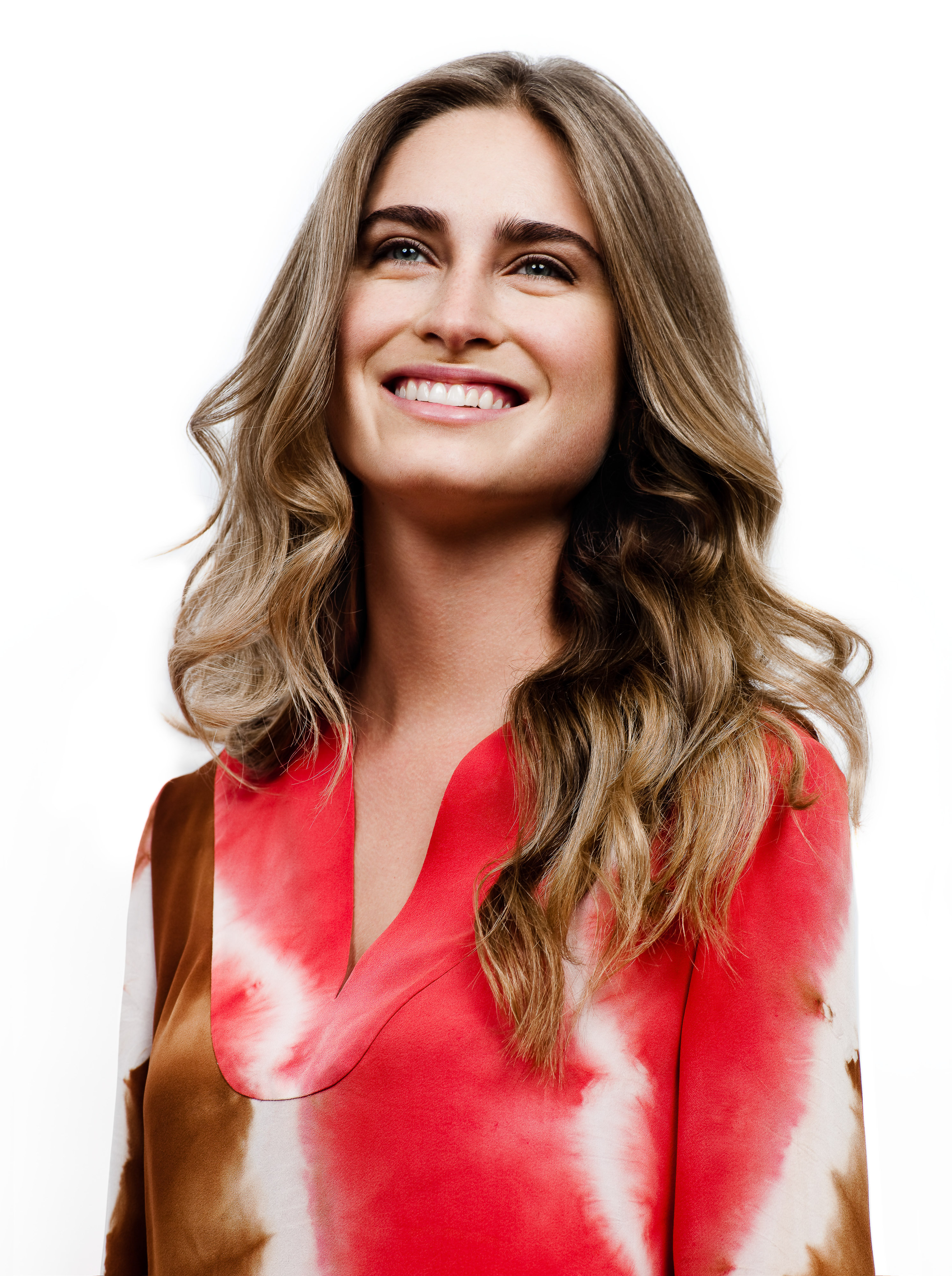 Portrait of a smiling adult woman with long blonde wavy hair, wearing a coral-colored long-sleeve top, set against a bright white backdrop.