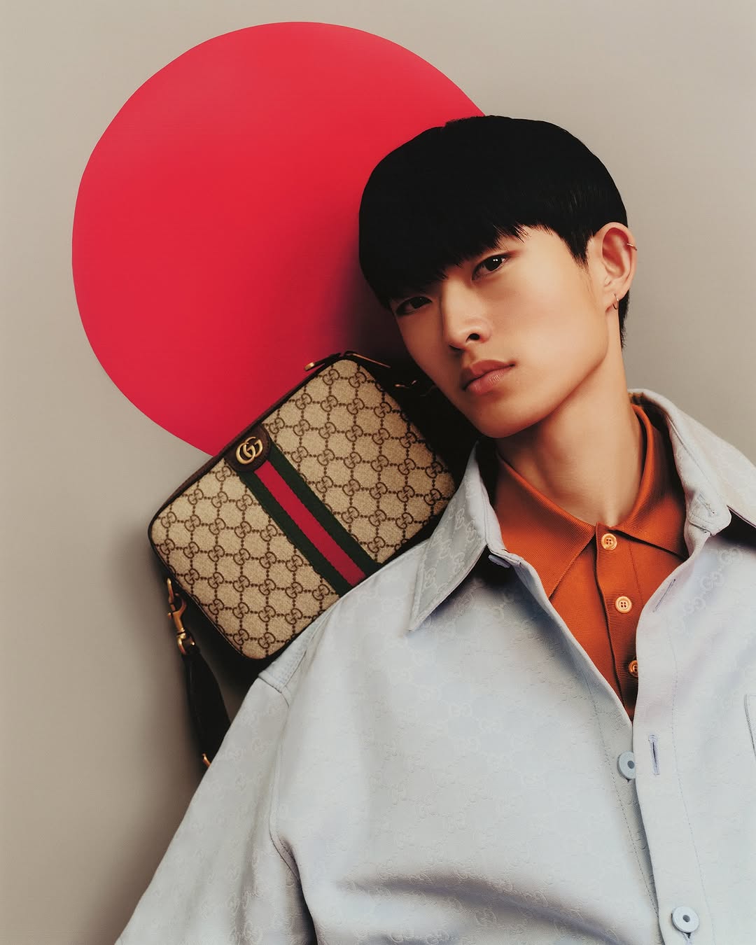 Asian male model poses with Gucci bag and a round red prop in a fashion studio shoot. Light grey backdrop, warm color scheme.