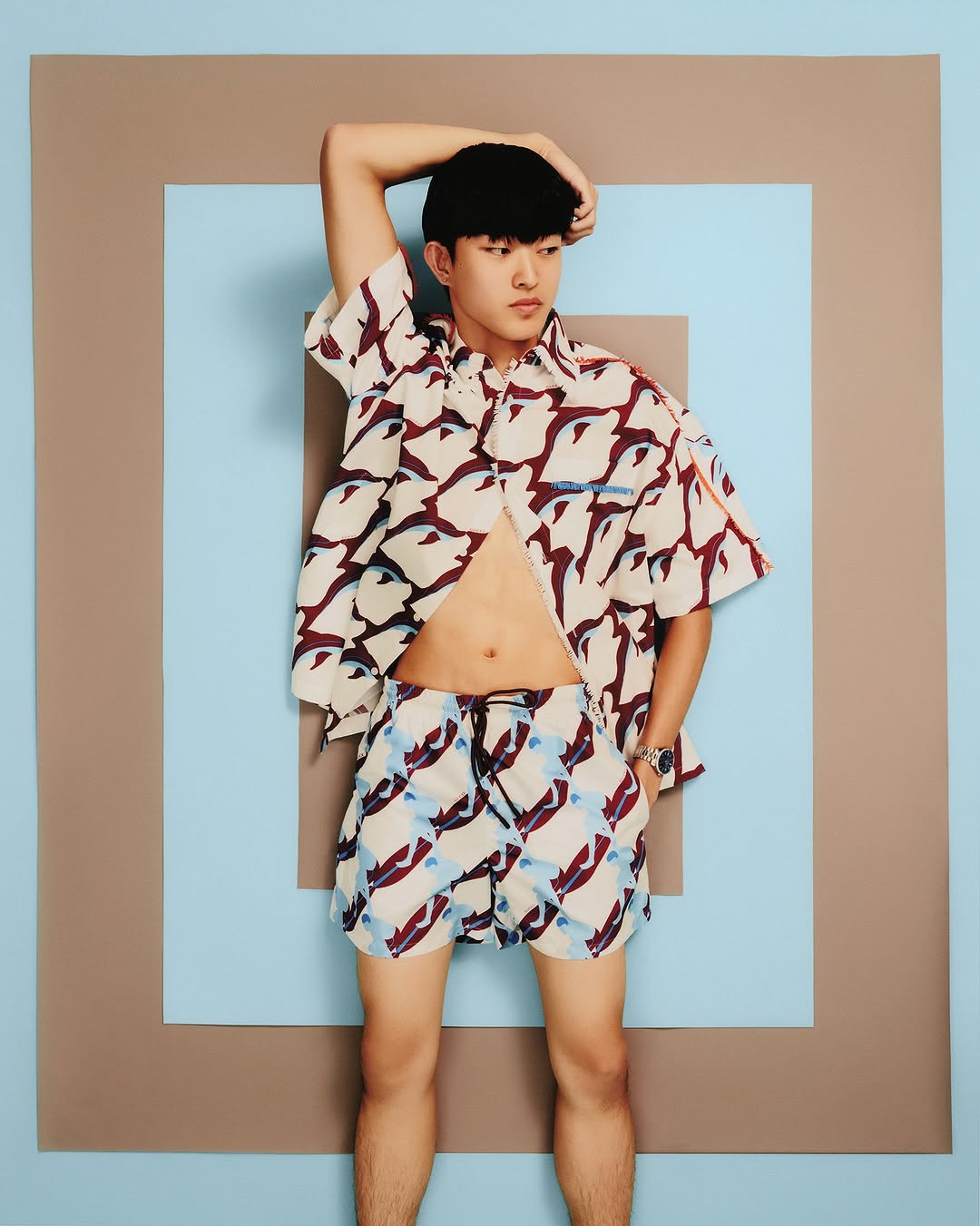 Young Asian man poses in a patterned shirt and shorts against a blue and tan square backdrop, showcasing summer fashion and style.