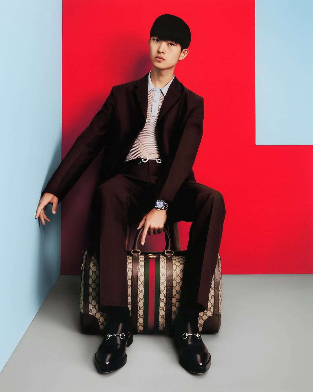 An Asian male fashion model poses seated on a Gucci bag, wearing a brown suit against a red and blue studio backdrop, looking at camera.