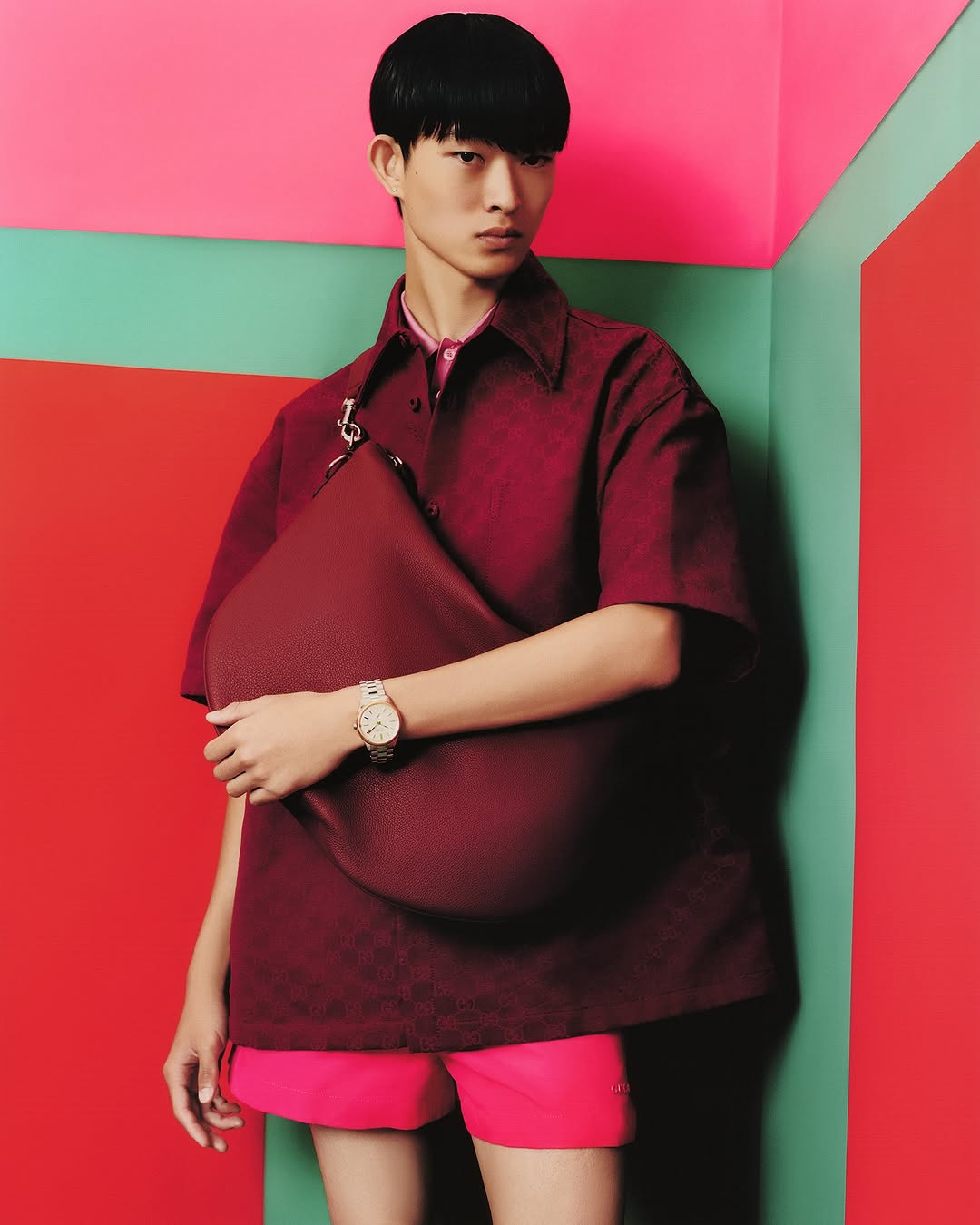 An Asian man poses in a red shirt and pink shorts, holding a matching bag, against a vibrant pink, red, and green wall.