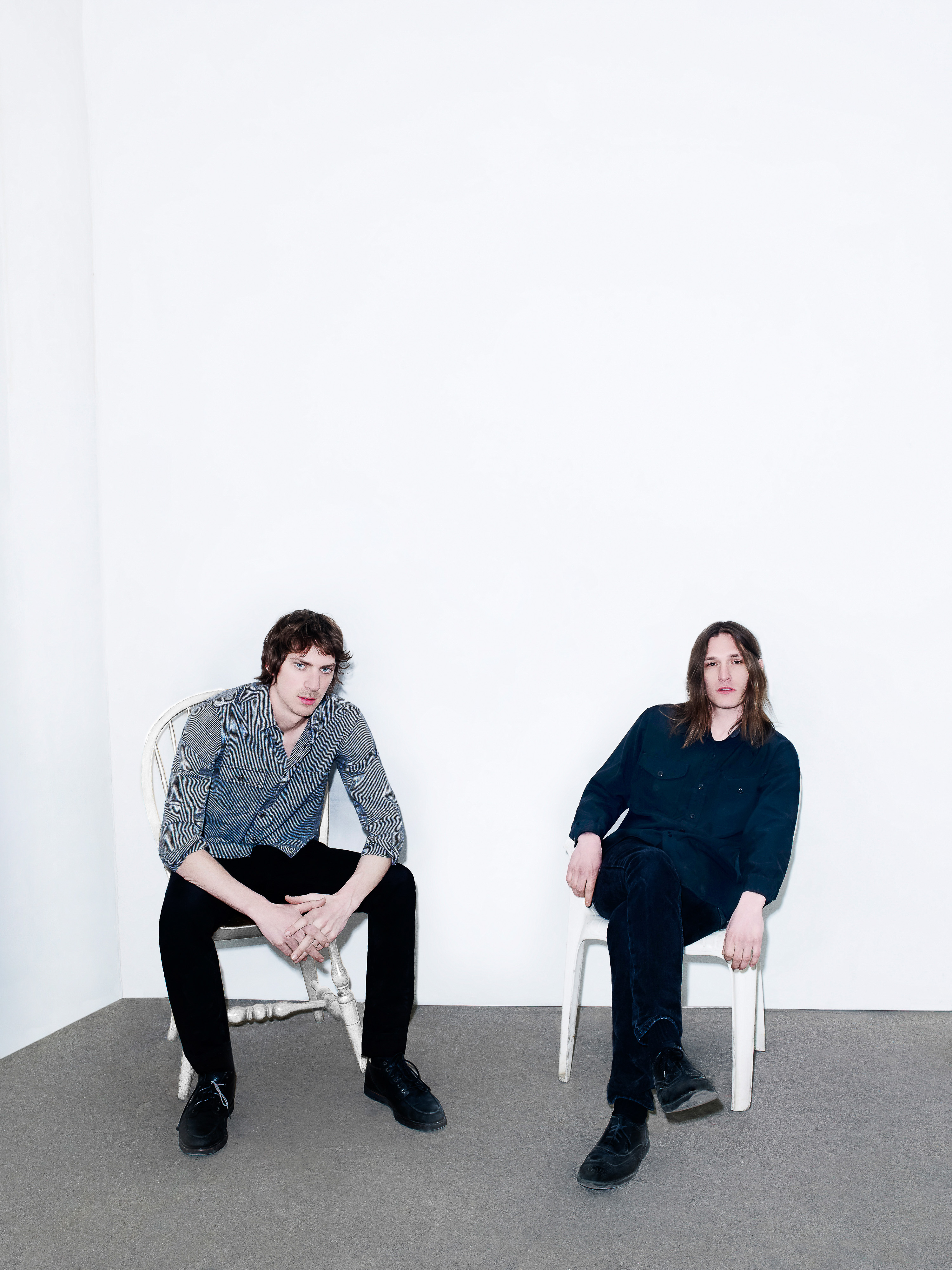 Two adult men sit on chairs in a bright white photo studio. One man wears a grey button-down shirt and black pants, the other in black.