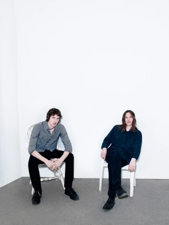 Two adult men sit on chairs in a bright white photo studio. One man wears a grey button-down shirt and black pants, the other in black.