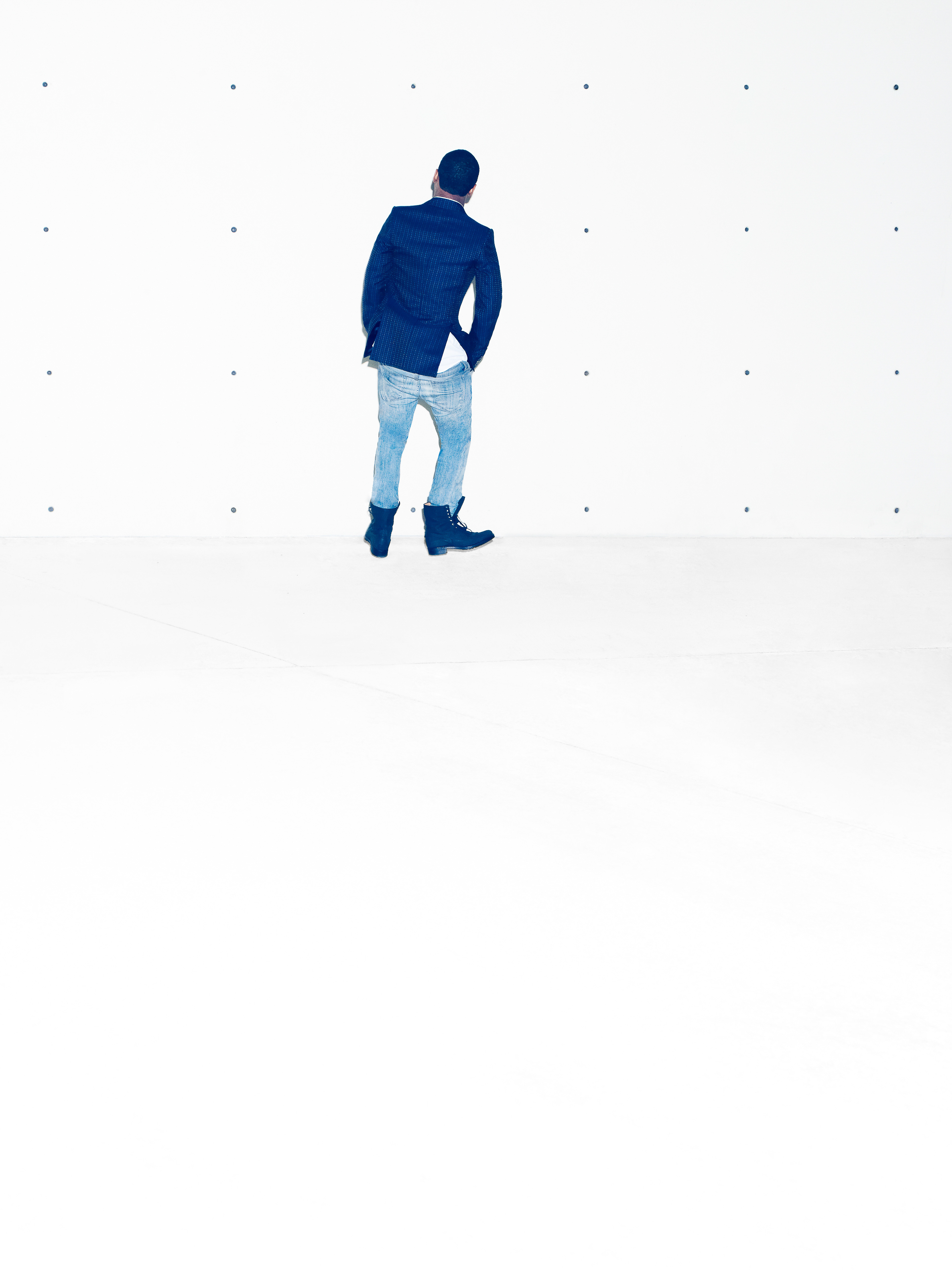 A person in a dark blue blazer and jeans stands in a white room with dark dots on the wall, viewed from the back.