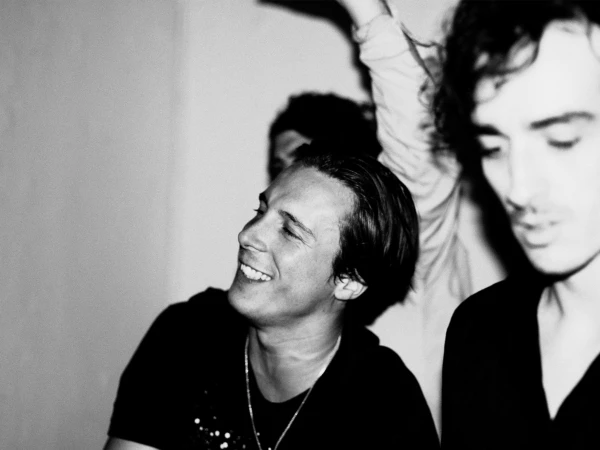 Black and white candid photograph of a smiling young man wearing a black t-shirt and thin chain necklace indoors.