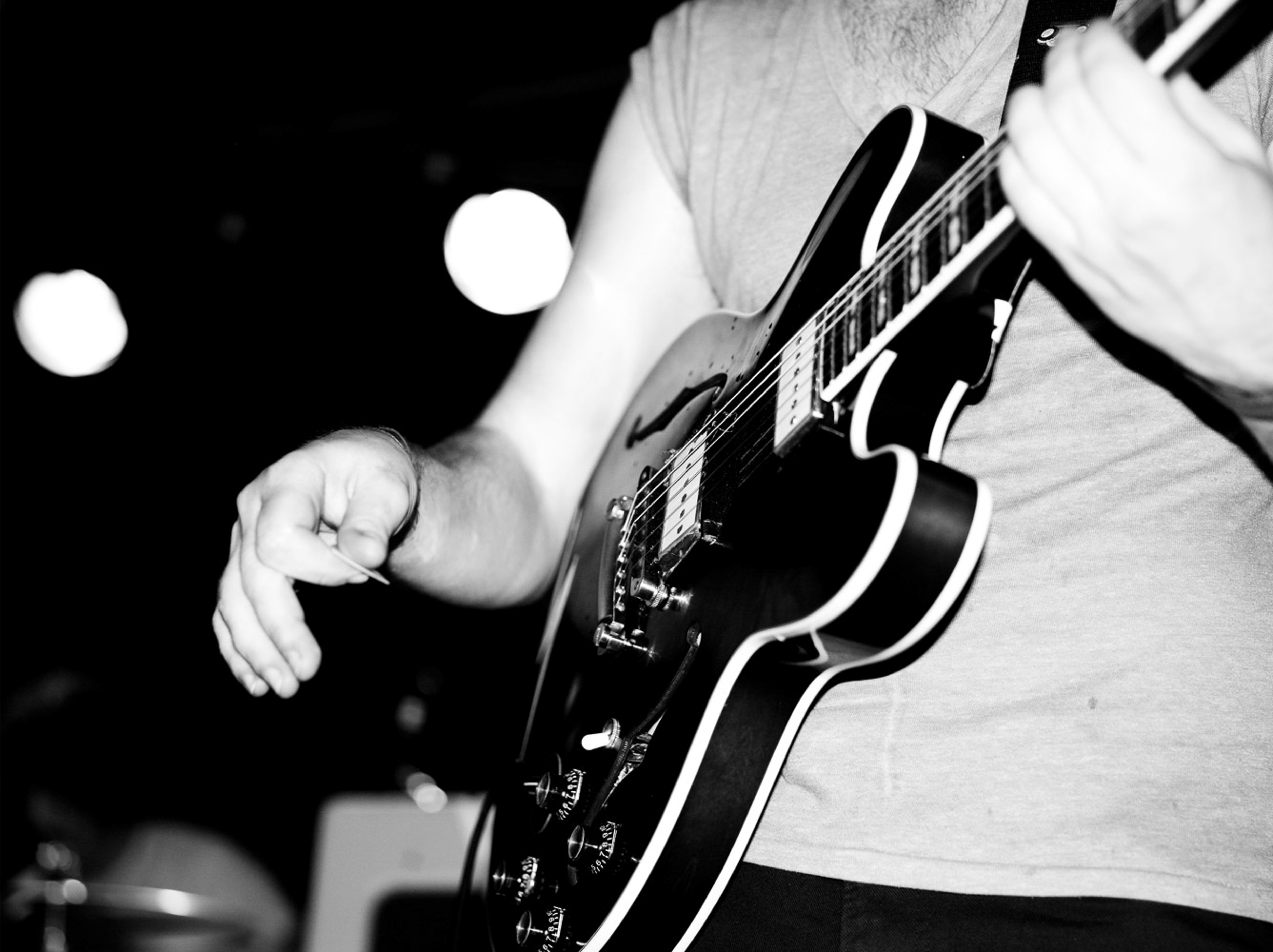 Black and white photo of a musician holding a guitar pick near an electric guitar, ready to strum, on a dark stage.