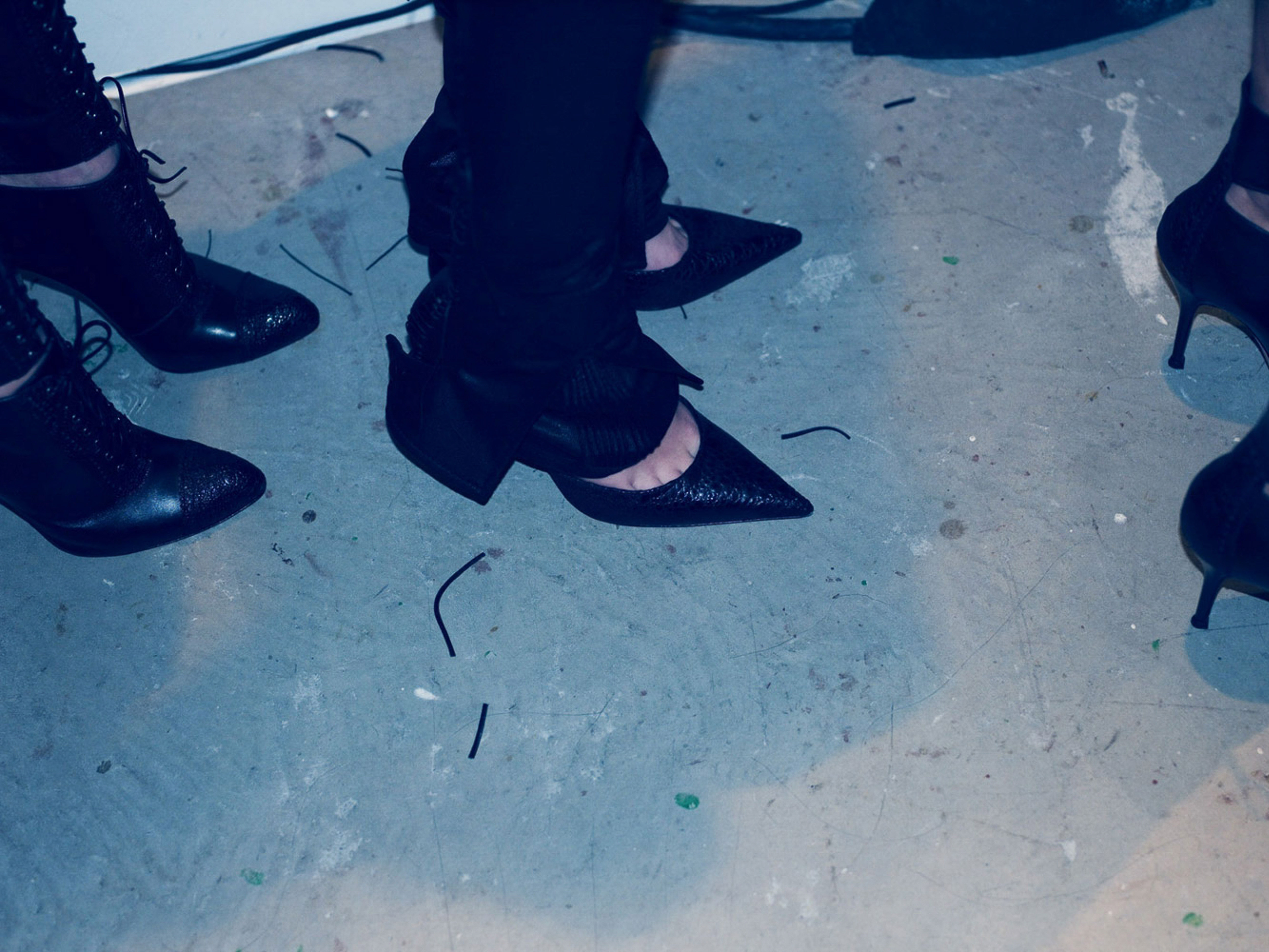 Close-up on four people wearing high-heeled black shoes indoors on a grey concrete floor with black strings scattered across the surface.