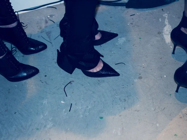 Close-up on four people wearing high-heeled black shoes indoors on a grey concrete floor with black strings scattered across the surface.