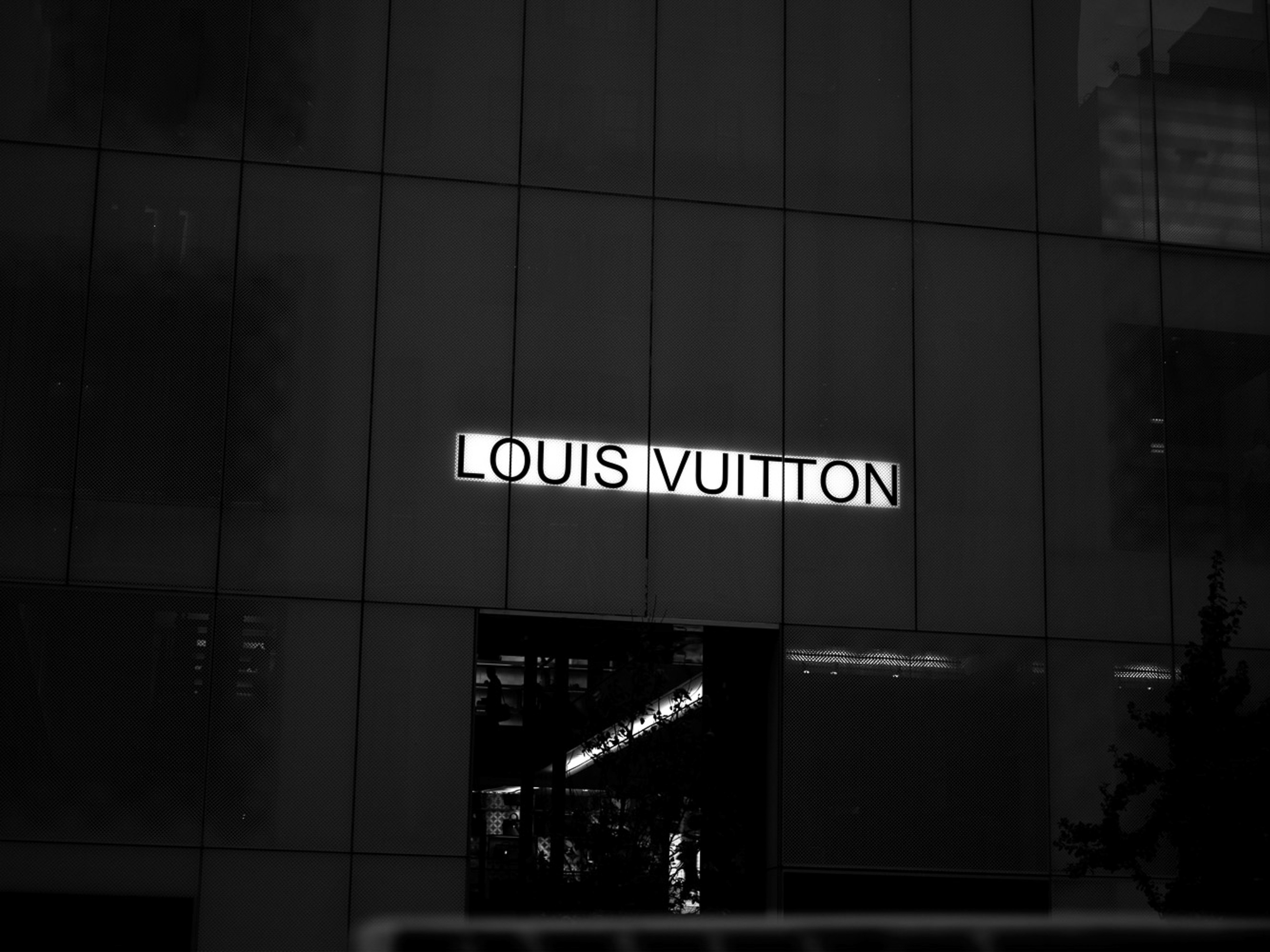 Louis Vuitton store facade at night, featuring a brightly lit logo against a dark tiled background in black and white photography.