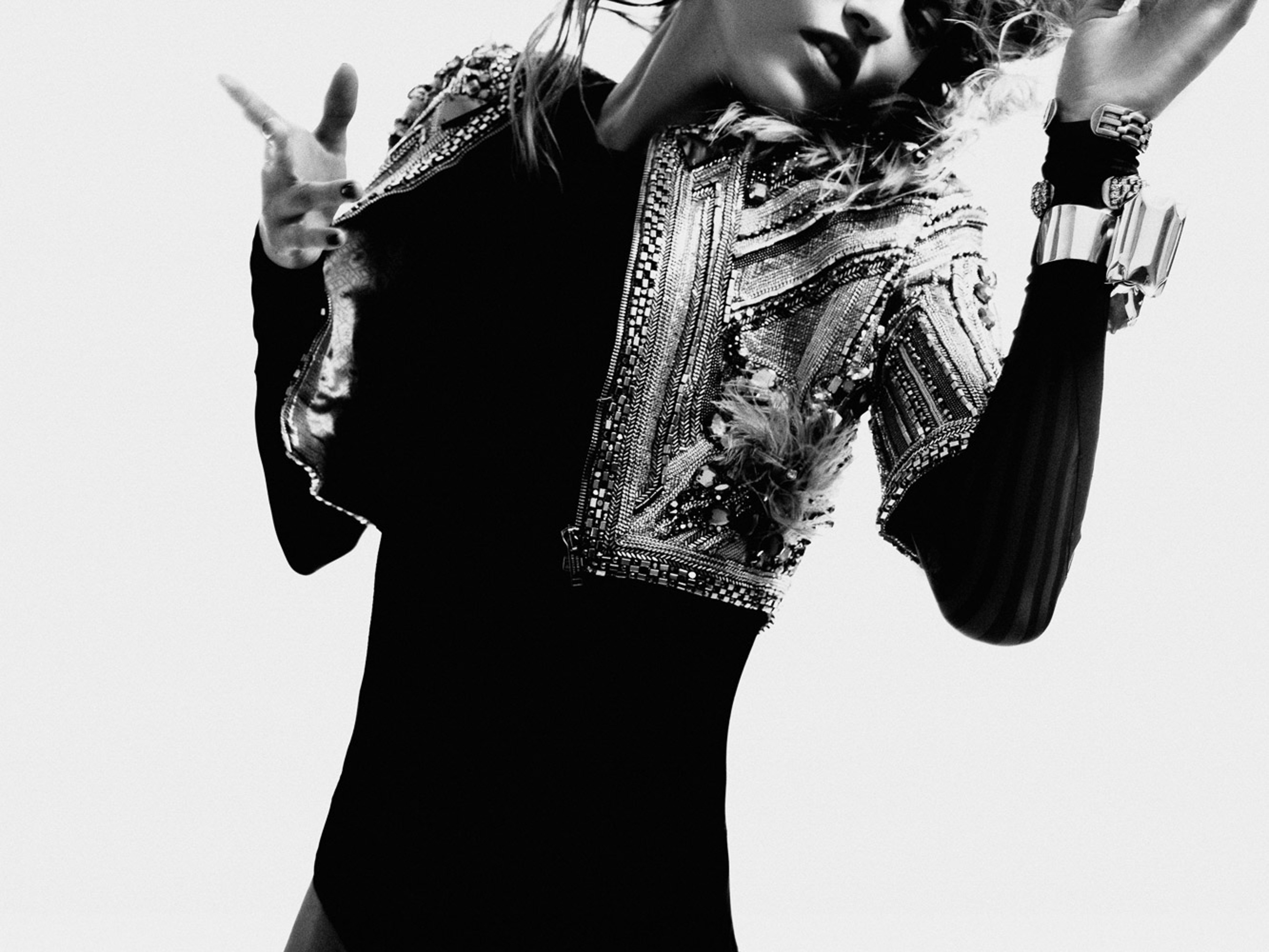 Woman in black long-sleeved dress and embellished jacket poses in a photo studio against a white backdrop with bracelets and rings.
