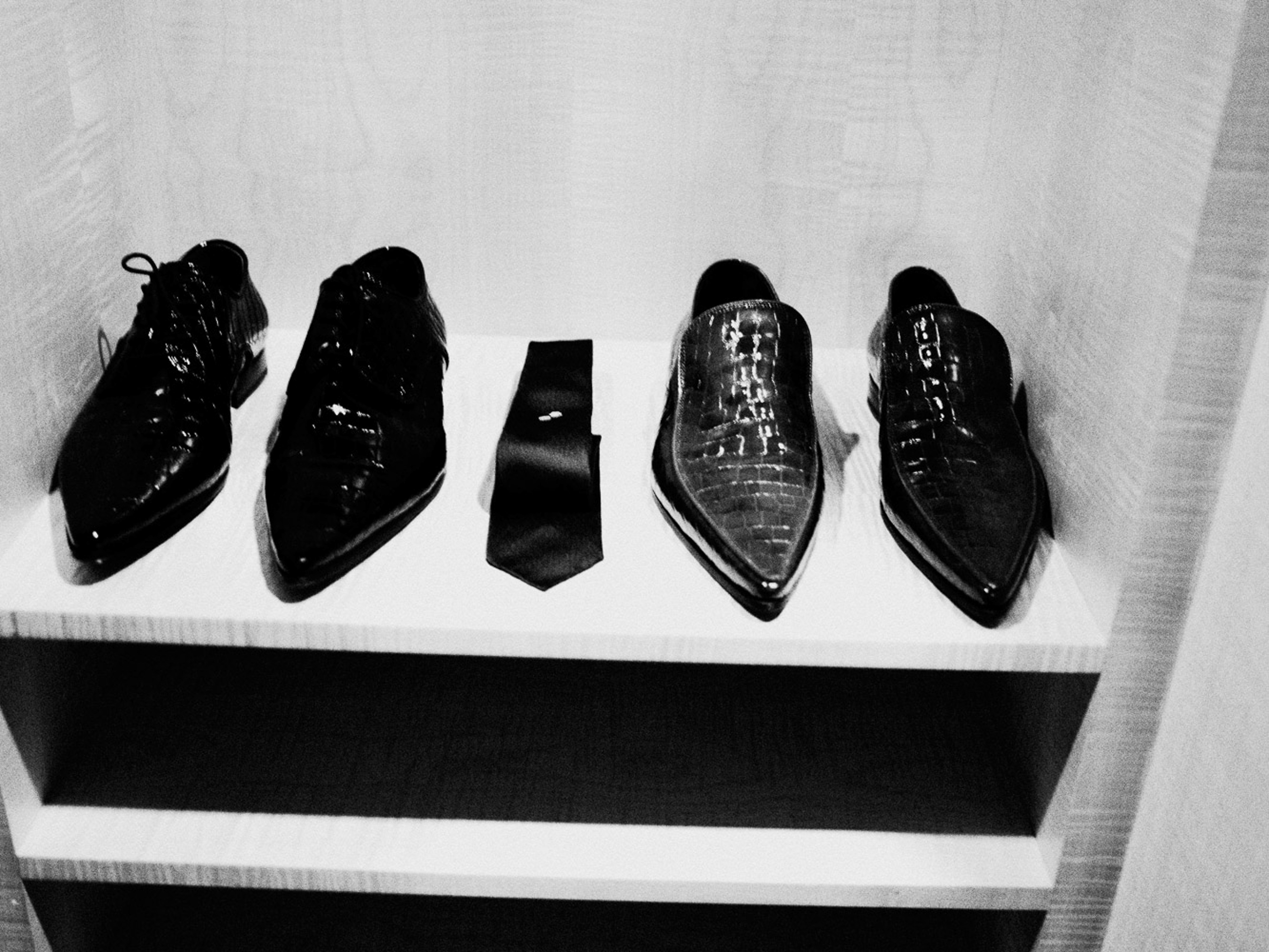 Black and white image of four shiny, pointed dress shoes and a tie on a shelf, offering a glimpse into men's fashion and style.
