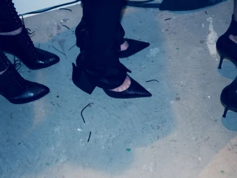 Models standing backstage wearing textured black pointed-toe heels, stiletto pumps, and lace-up boots on a concrete studio floor.