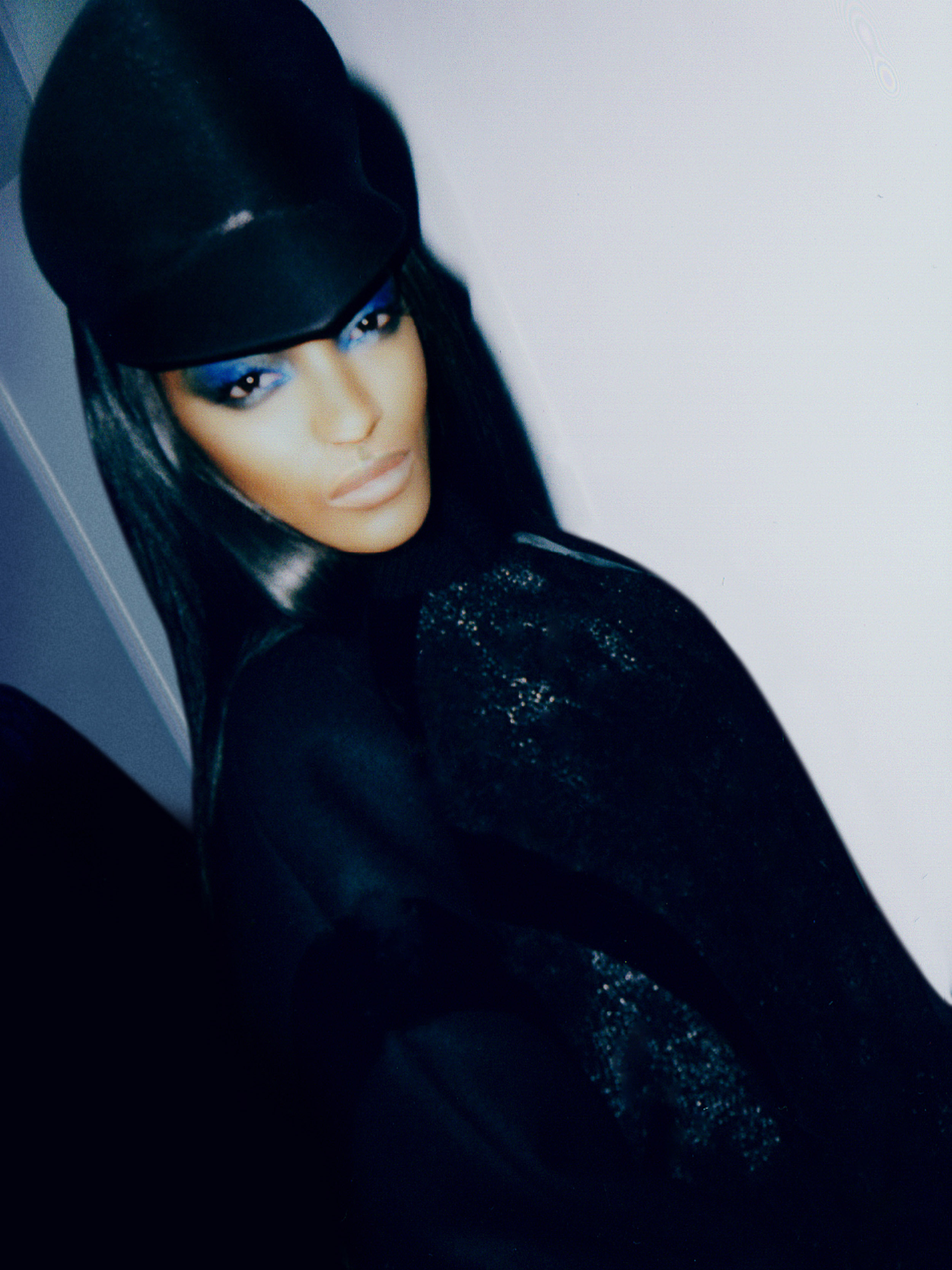 Close-up portrait of a stylish adult female with dark hair, wearing a black hat and black clothing with blue eyeshadow makeup.