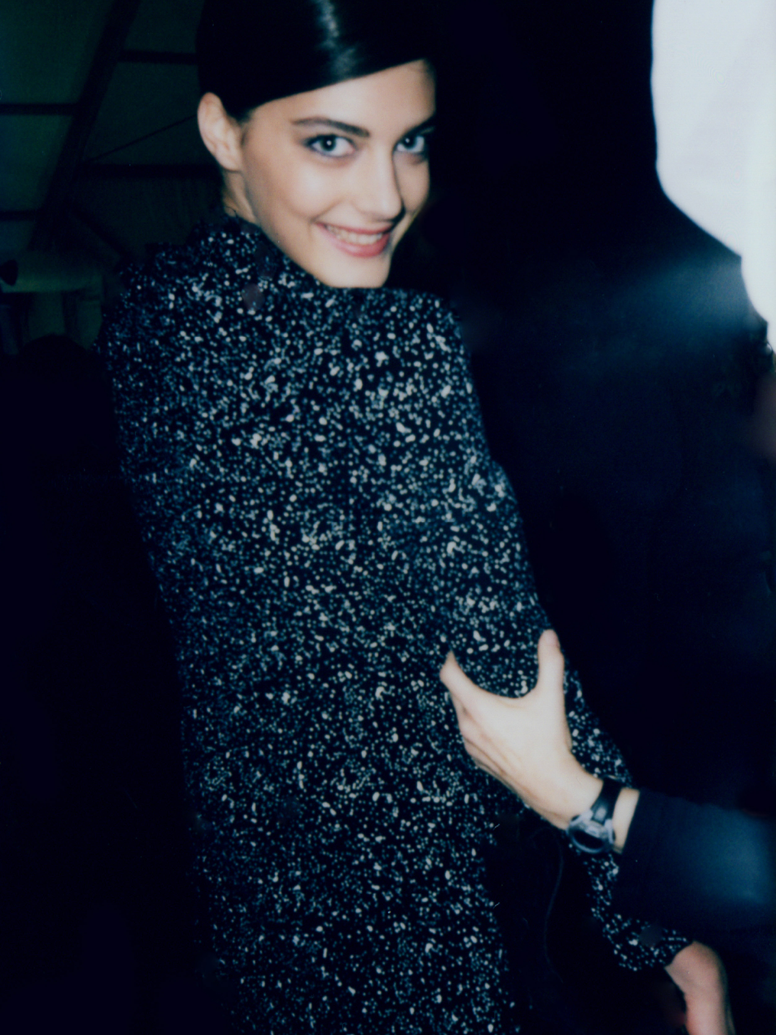 A smiling adult woman with dark hair styled back, wearing a patterned black-and-white coat, stands indoors in dark lighting with a helping hand.