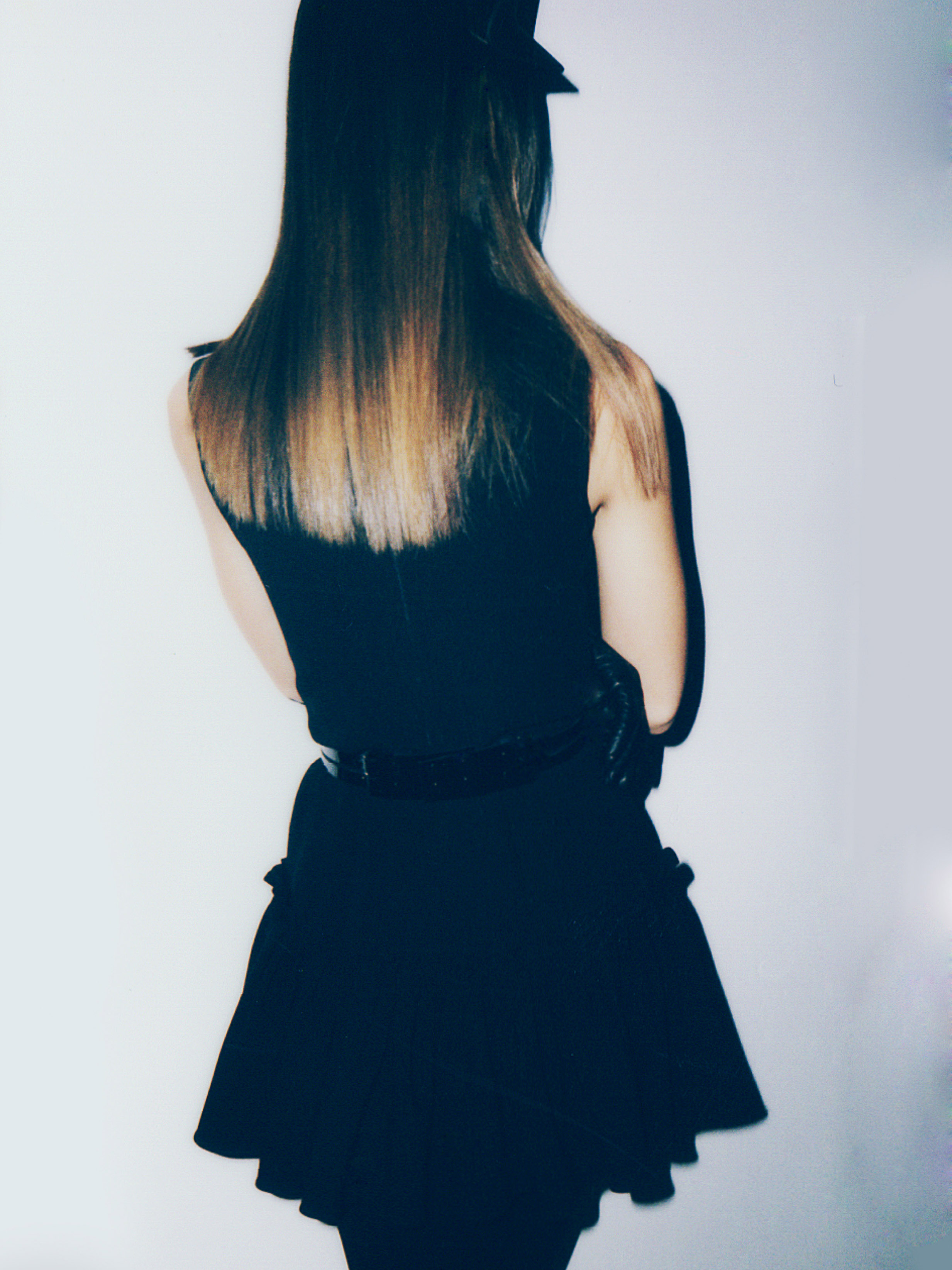 Back view of a person with long brown hair wearing a black dress, belt, gloves and hat standing in front of a white background.
