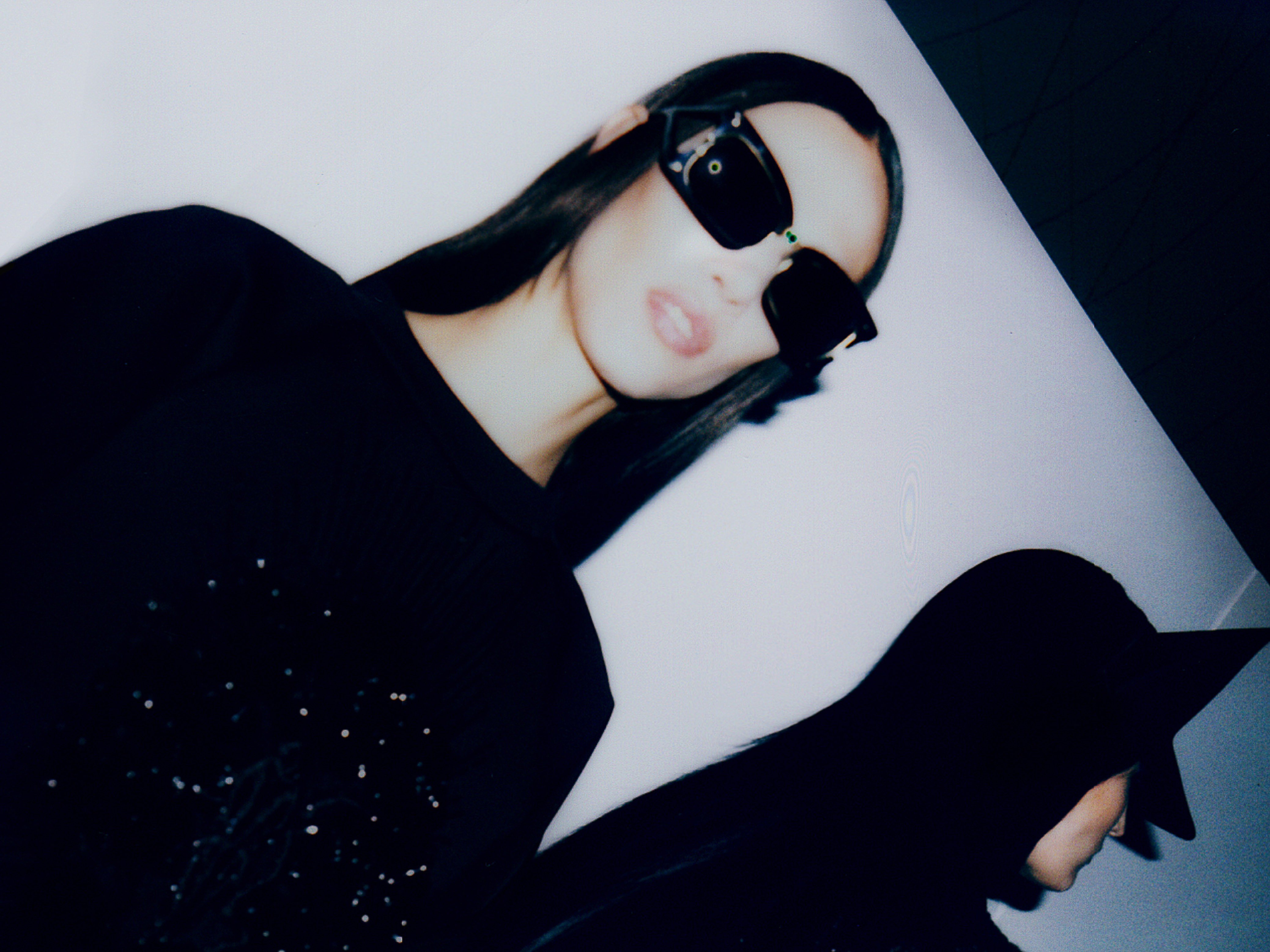 Two women with sunglasses and black tops pose in a studio against a white wall, creating a stark, stylish image with strong contrasts.