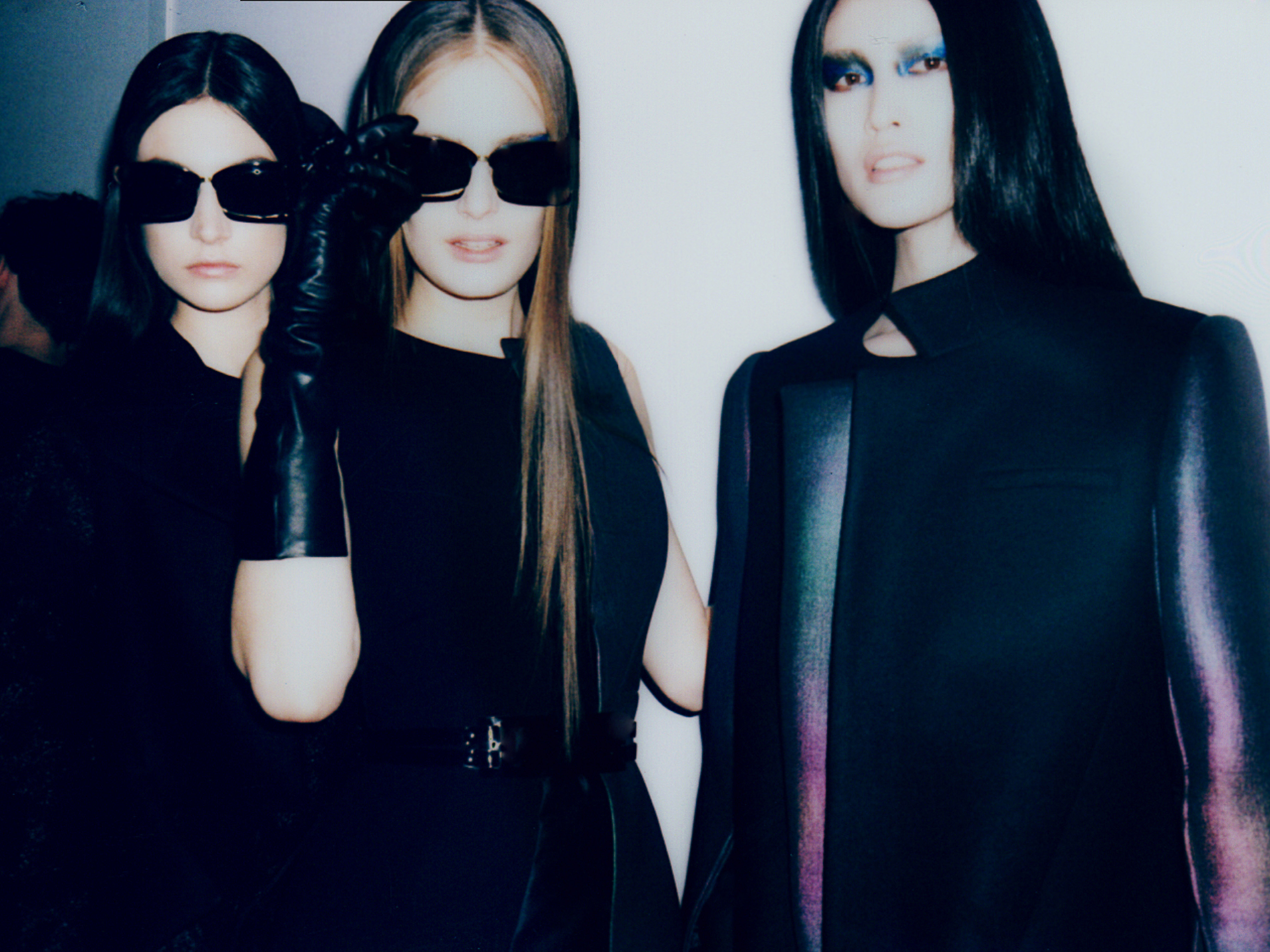 Three fashion models pose in black clothing and sunglasses against a white wall, exuding a dark, avant-garde aesthetic. One model wears gloves.