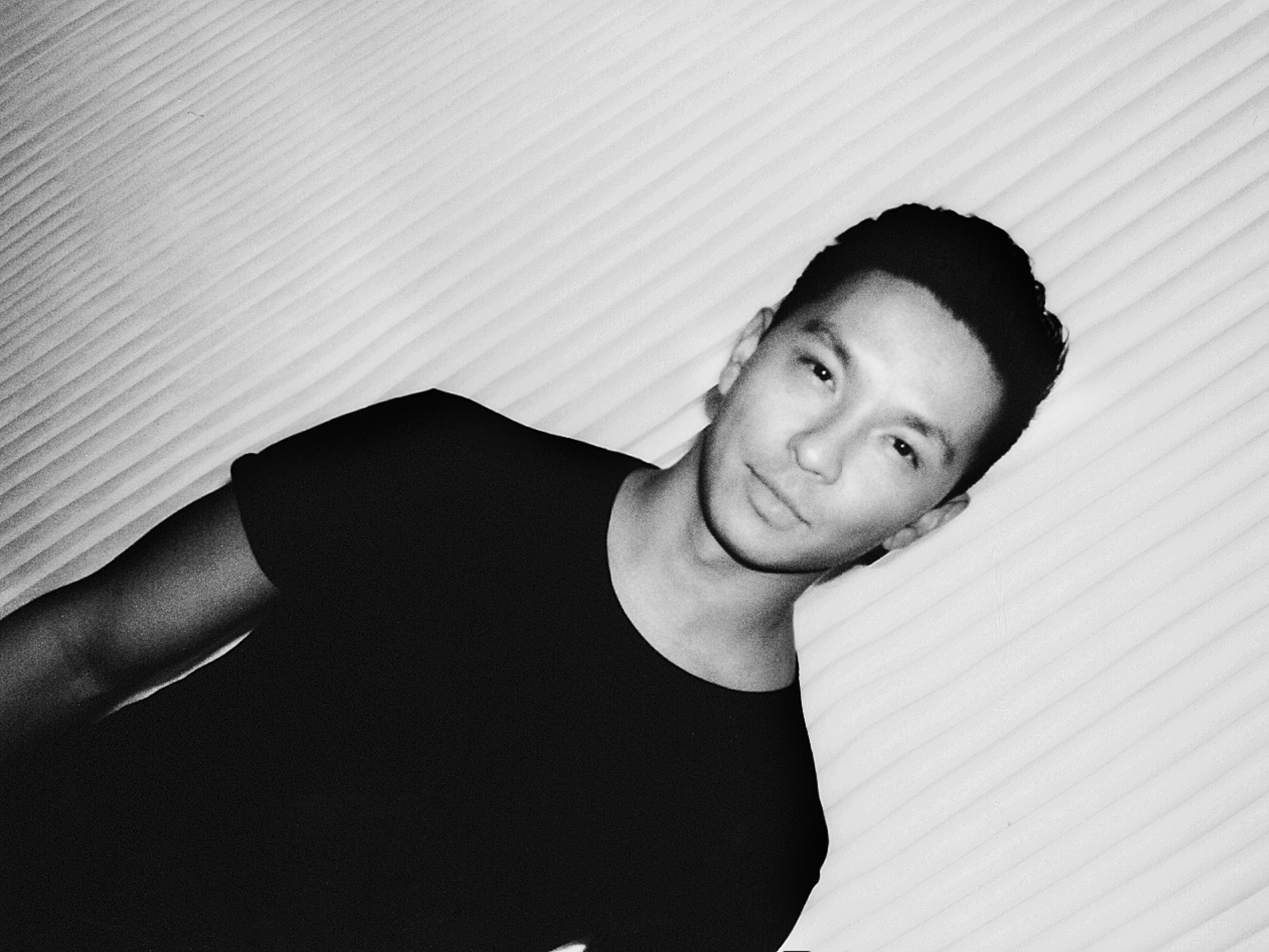 Monochrome portrait of a young adult male with dark hair, smiling subtly in front of a white corrugated wall.