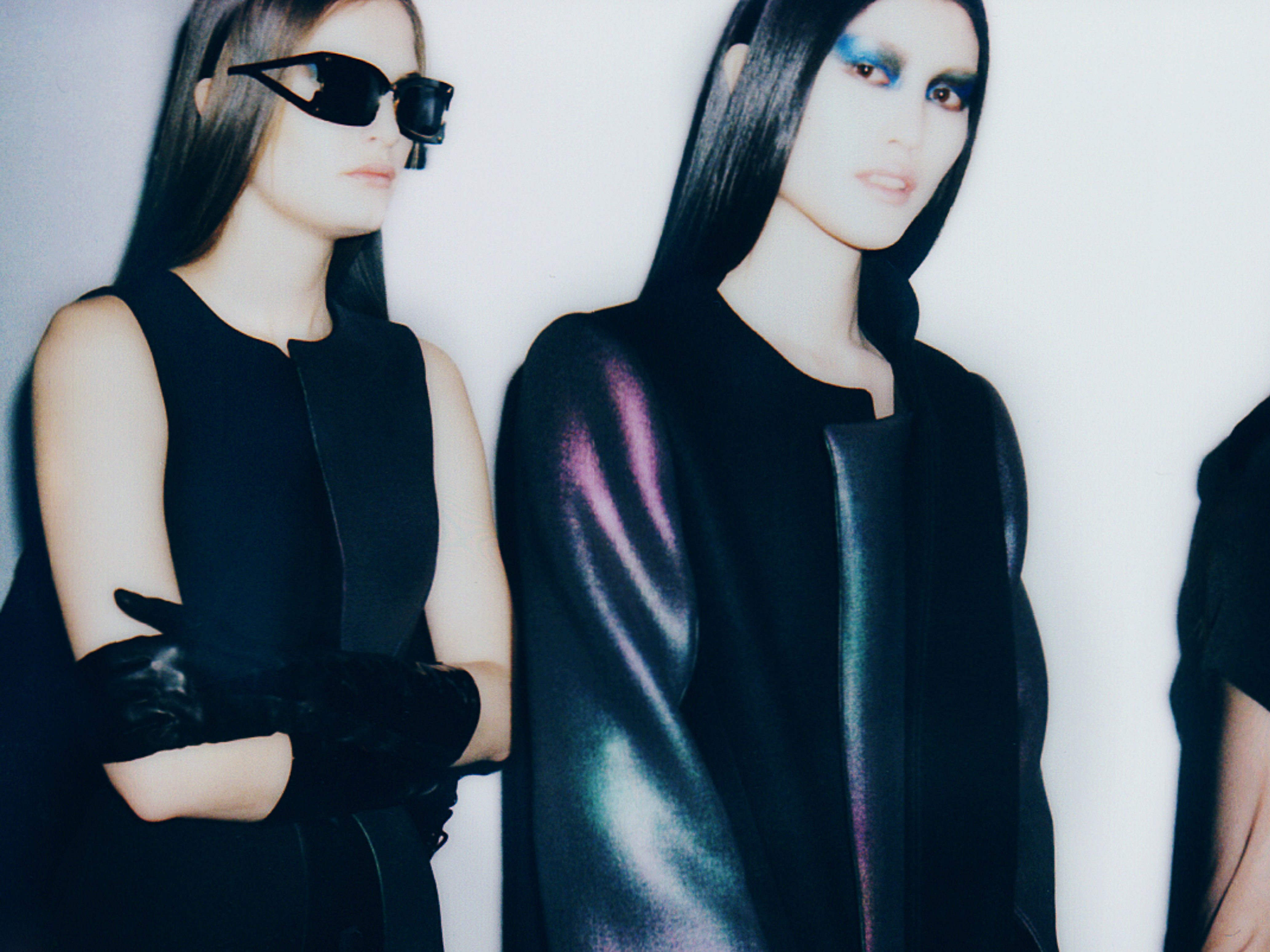 Two fashion models in black dresses and gloves pose in a studio, one wears sunglasses and the other dramatic makeup, against a white backdrop.