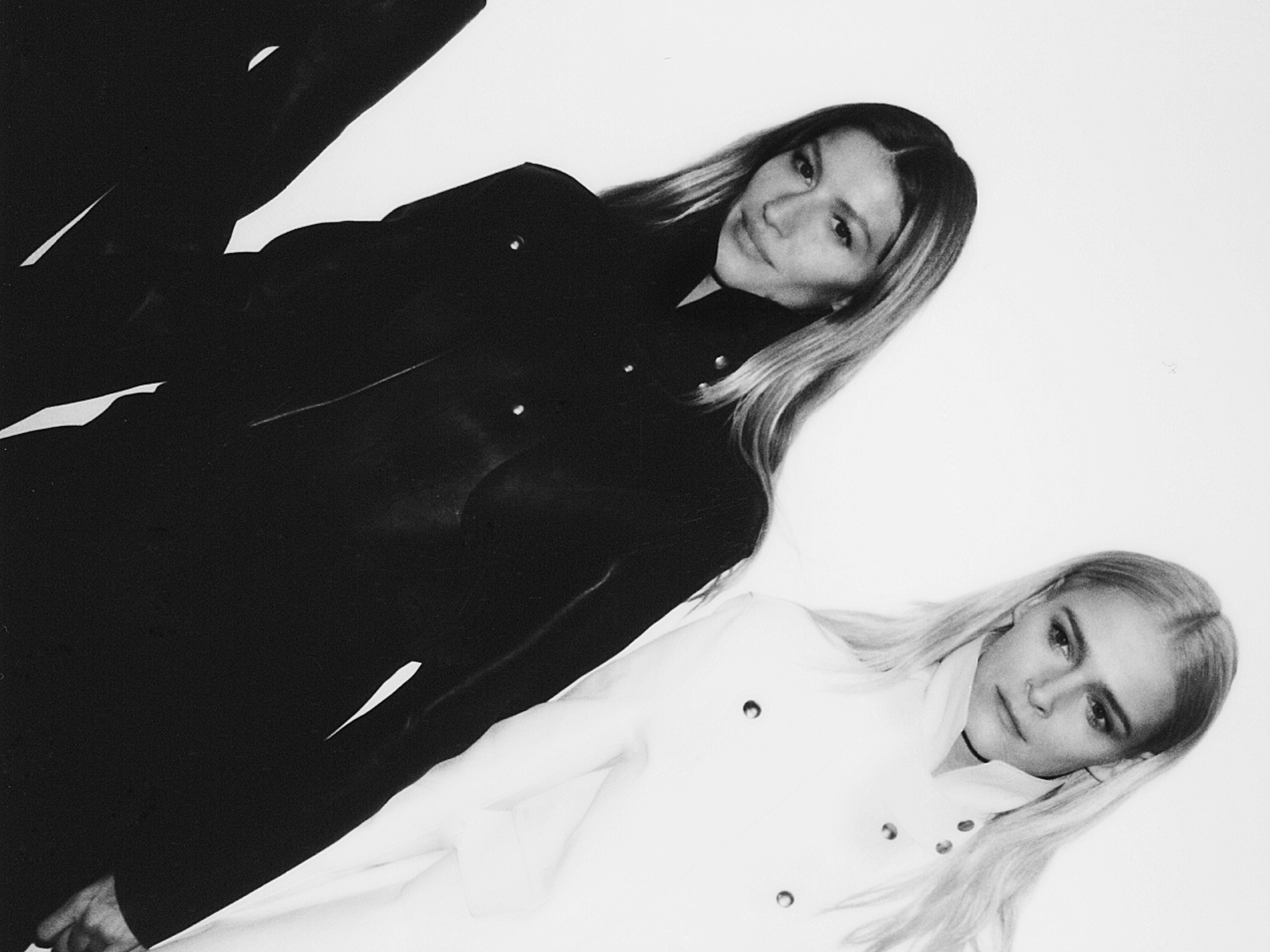 Black and white studio shot of two adult women with long blonde hair wearing similar jackets in complementary colors.