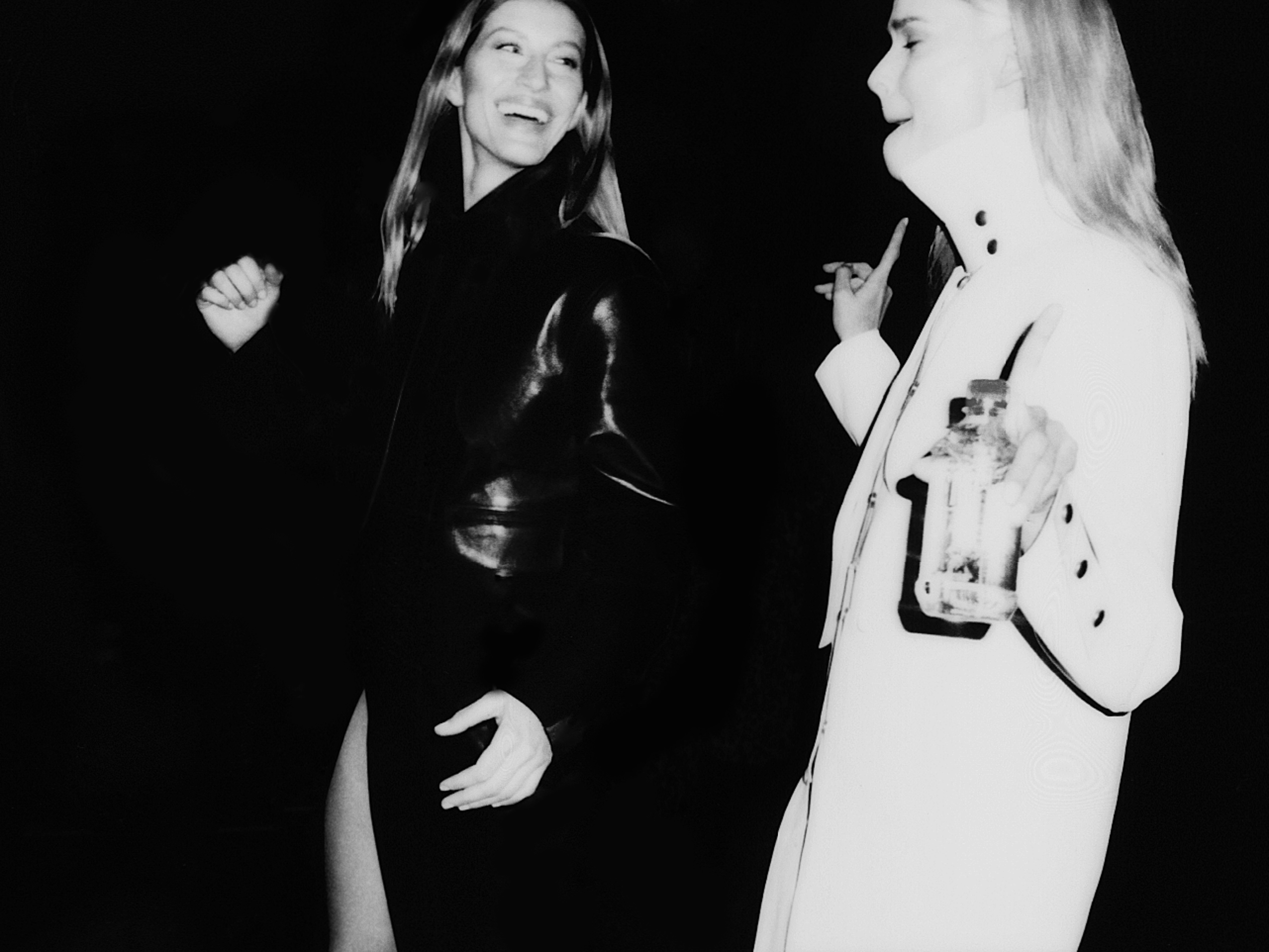 Two adult women in black-and-white image; one in a dark dress laughing, another in a light coat holding a bottle against a dark backdrop.
