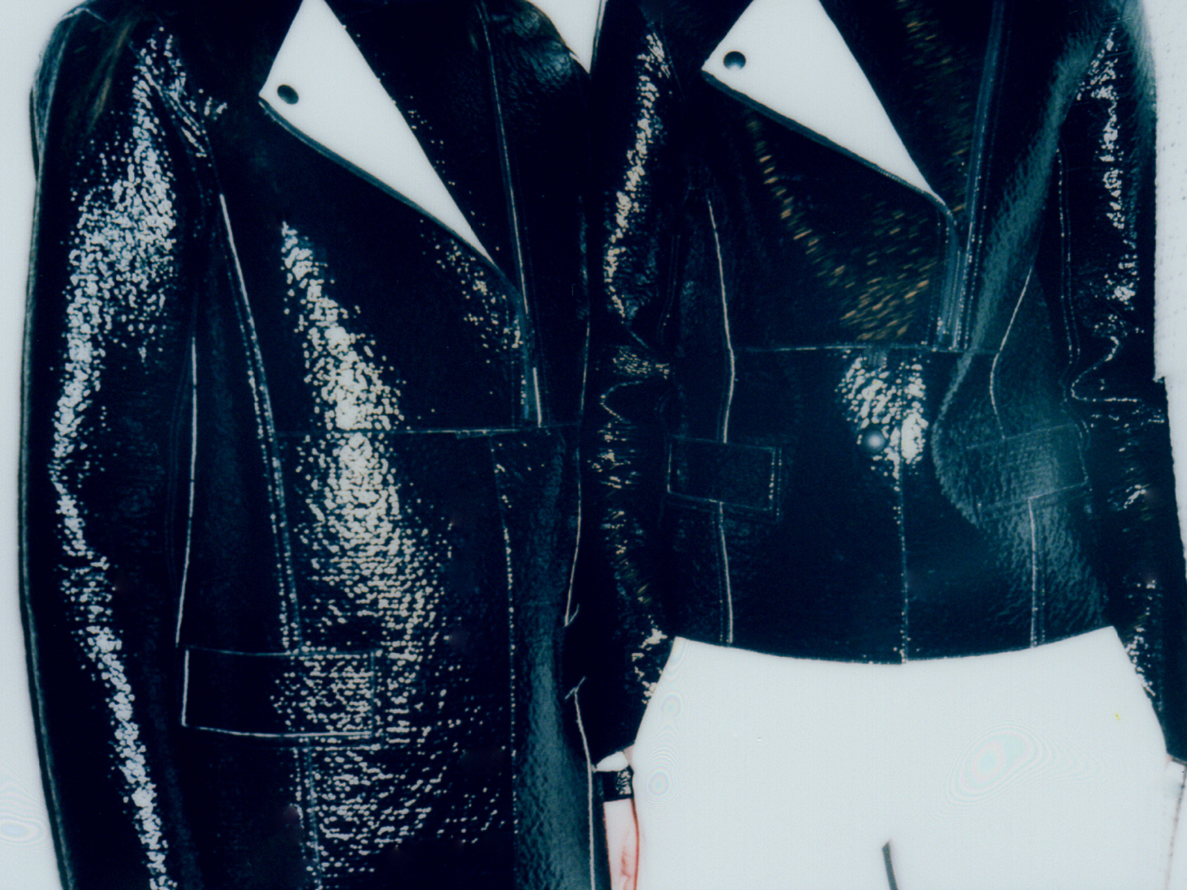 Two figures wearing shiny black patent leather moto jackets with white undershirts stand close together in a fashion forward portrait.
