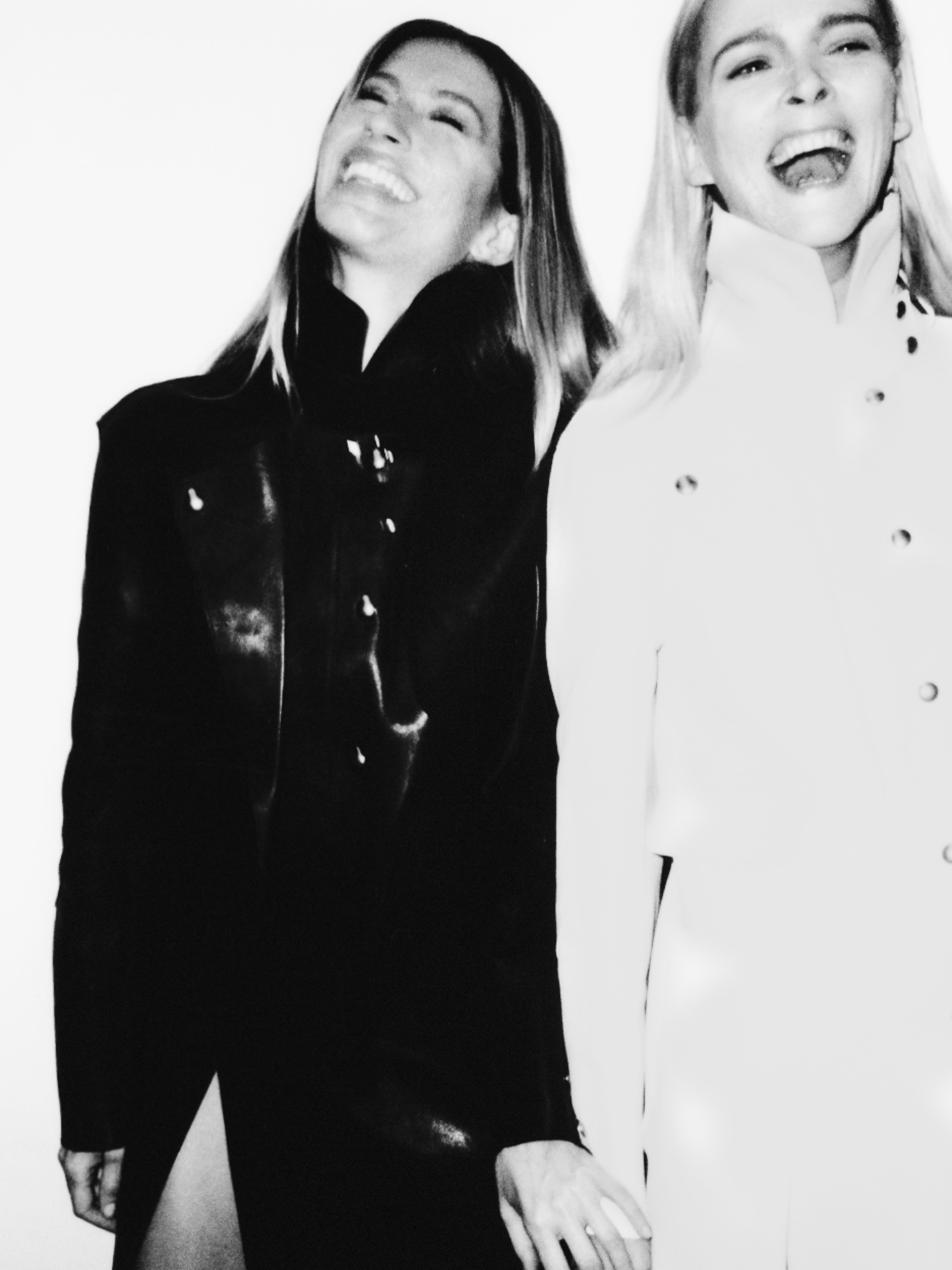 Two adult women laugh in a black and white close-up portrait. One wears a dark, button-down jacket, the other wears a similar white jacket.