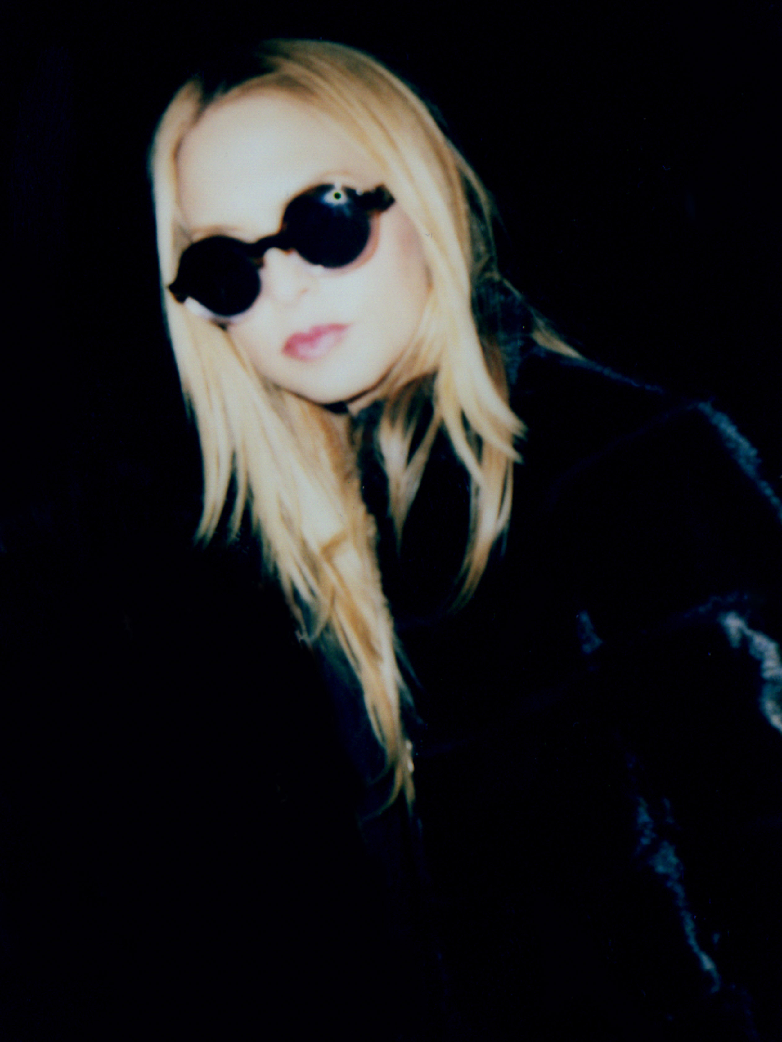 Stylish woman with blonde hair and round sunglasses poses in a dark indoor setting, wearing a black coat, creating a mysterious and fashionable look.